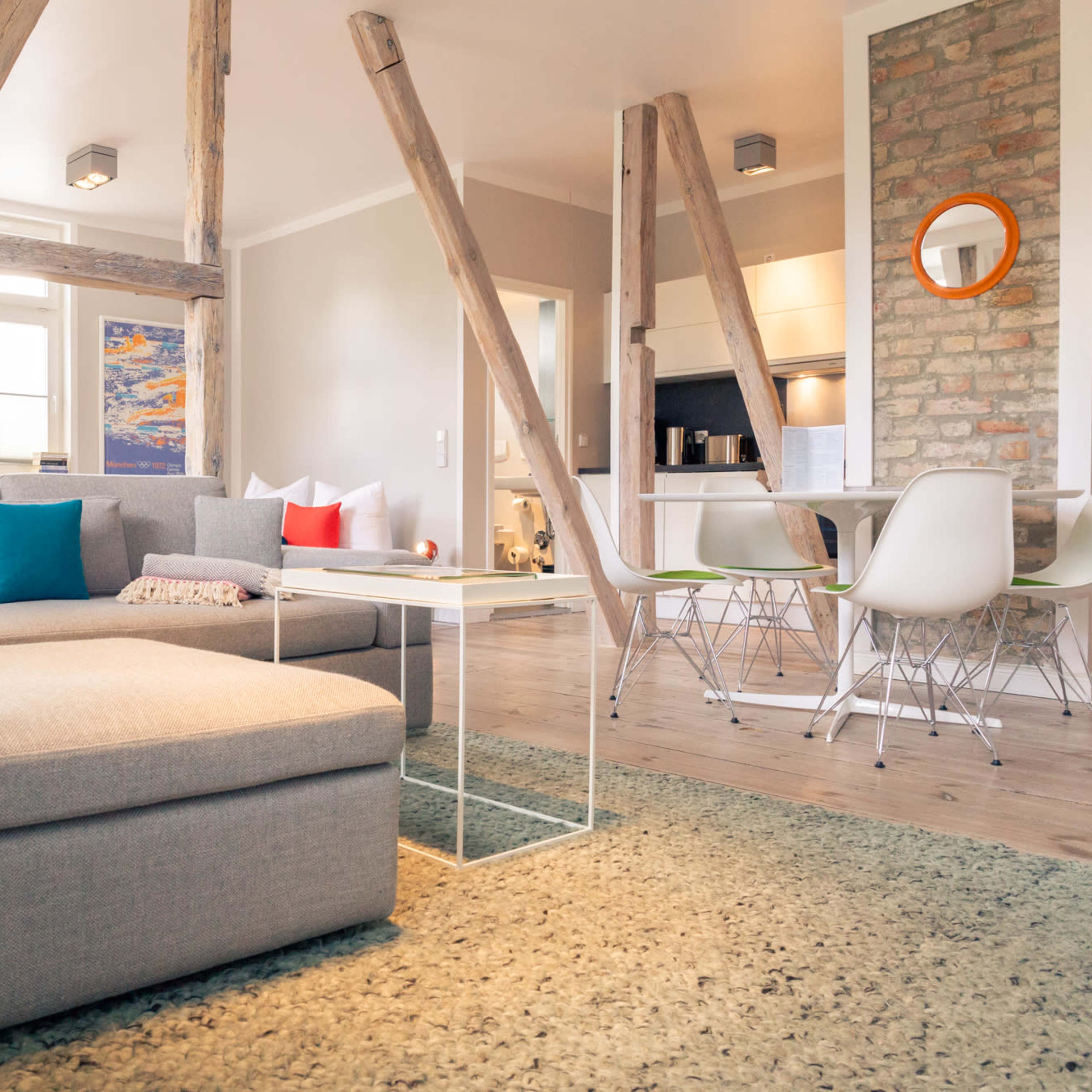 A modern living room with a gray sectional sofa, a coffee table, and an open kitchen area, featuring wooden beams and a textured rug.