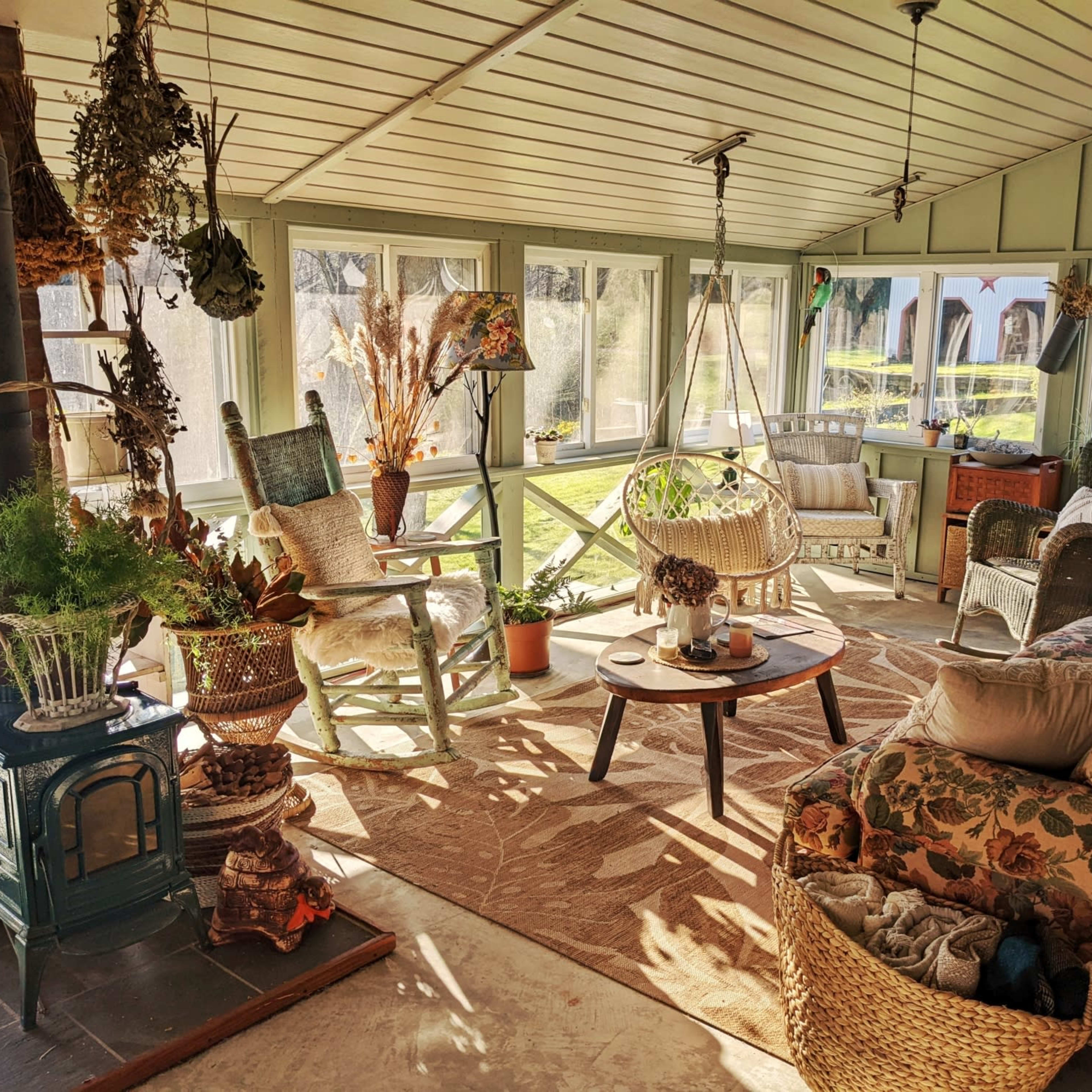 A sunlit sunroom furnished with a floral-patterned sofa, wicker chairs, a wooden table, and various plants.