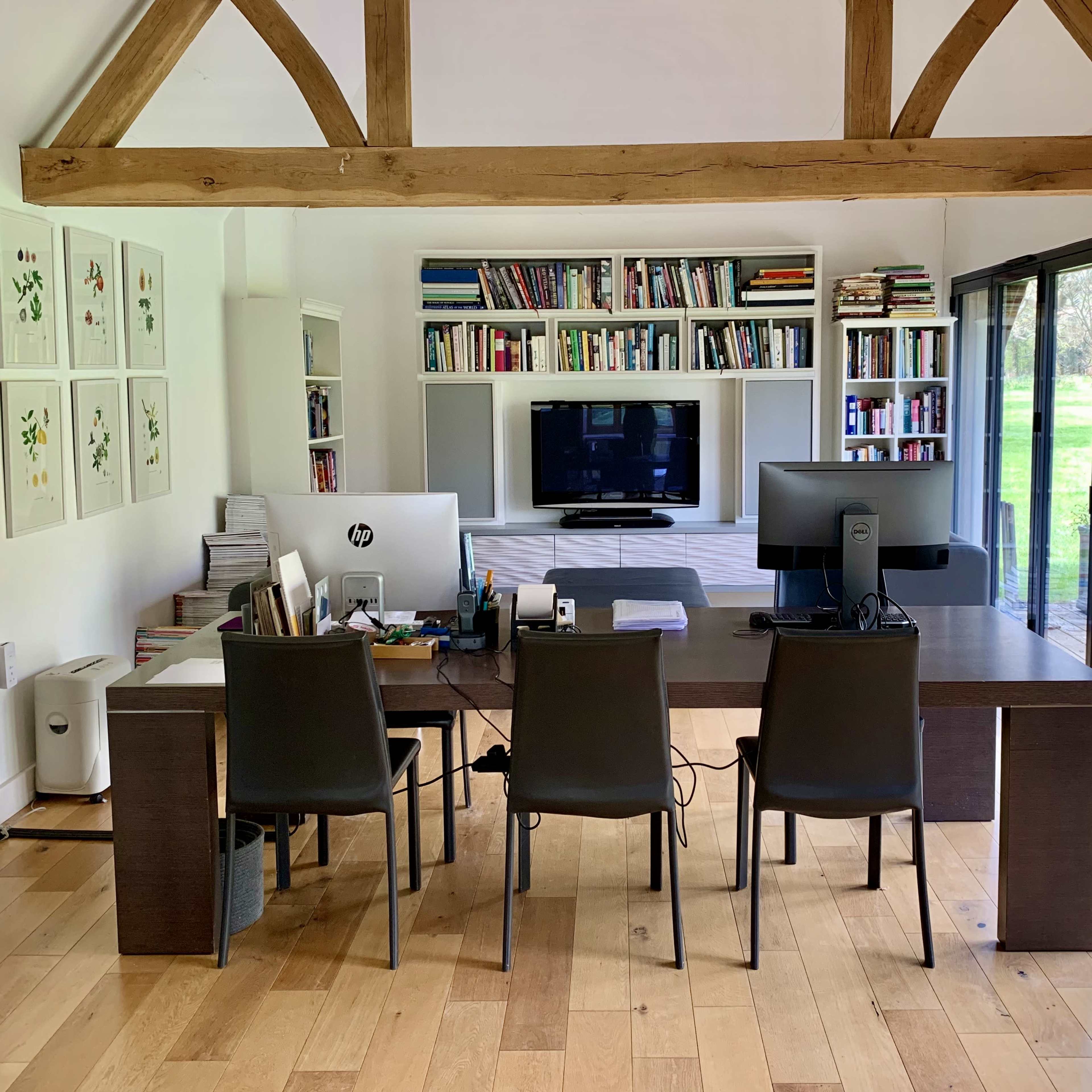 A modern office space with a large wooden desk, three black chairs, and bookshelves filled with books, facing a wall-mounted television and large windows that overlook a garden.