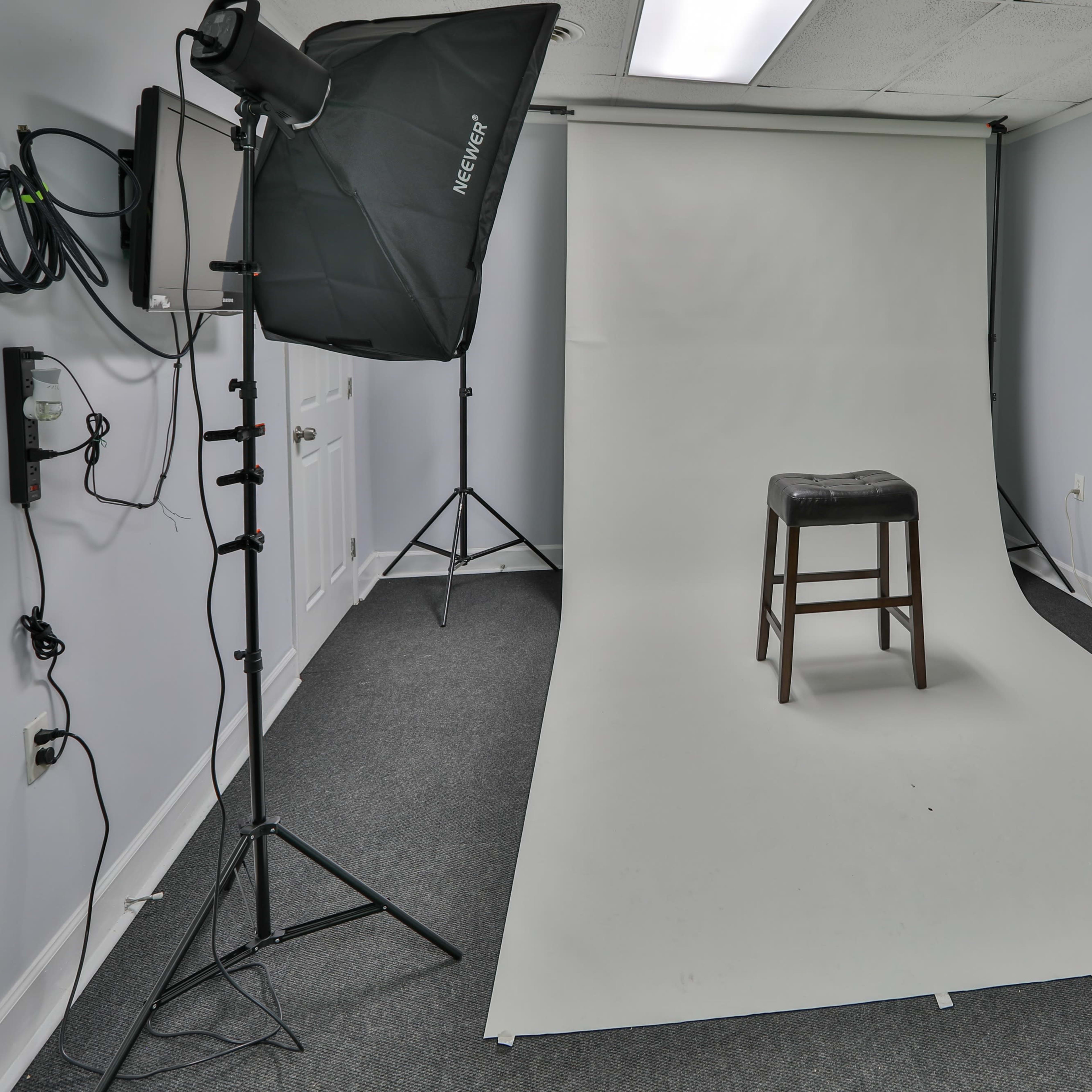 A photography studio features a bare white backdrop with a single wooden stool positioned in the center.