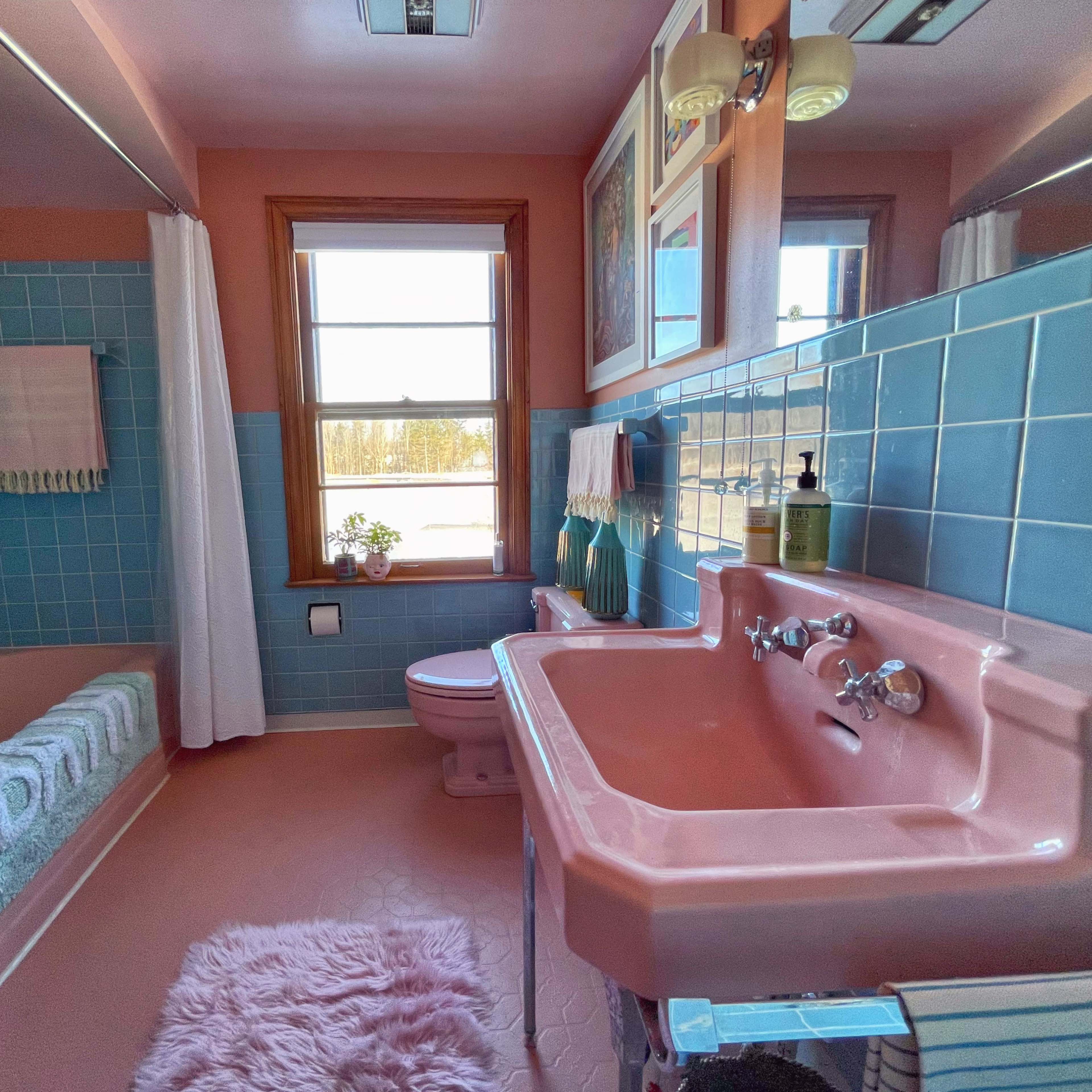 The bathroom features a pink sink and bathtub surrounded by blue tile and large windows letting in natural light.