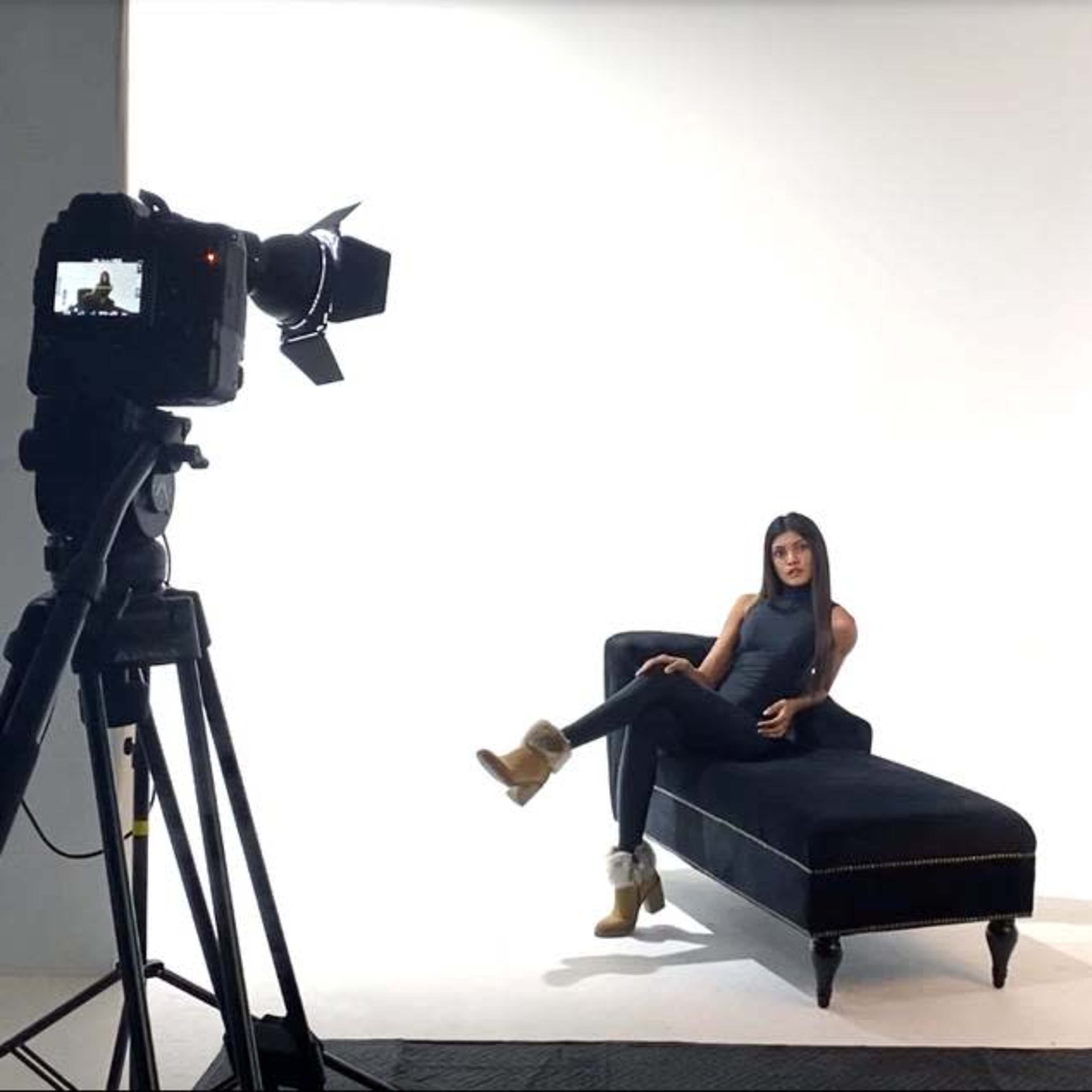 A woman sits on a black chaise lounge in a studio setting with a camera and lighting equipment positioned nearby.