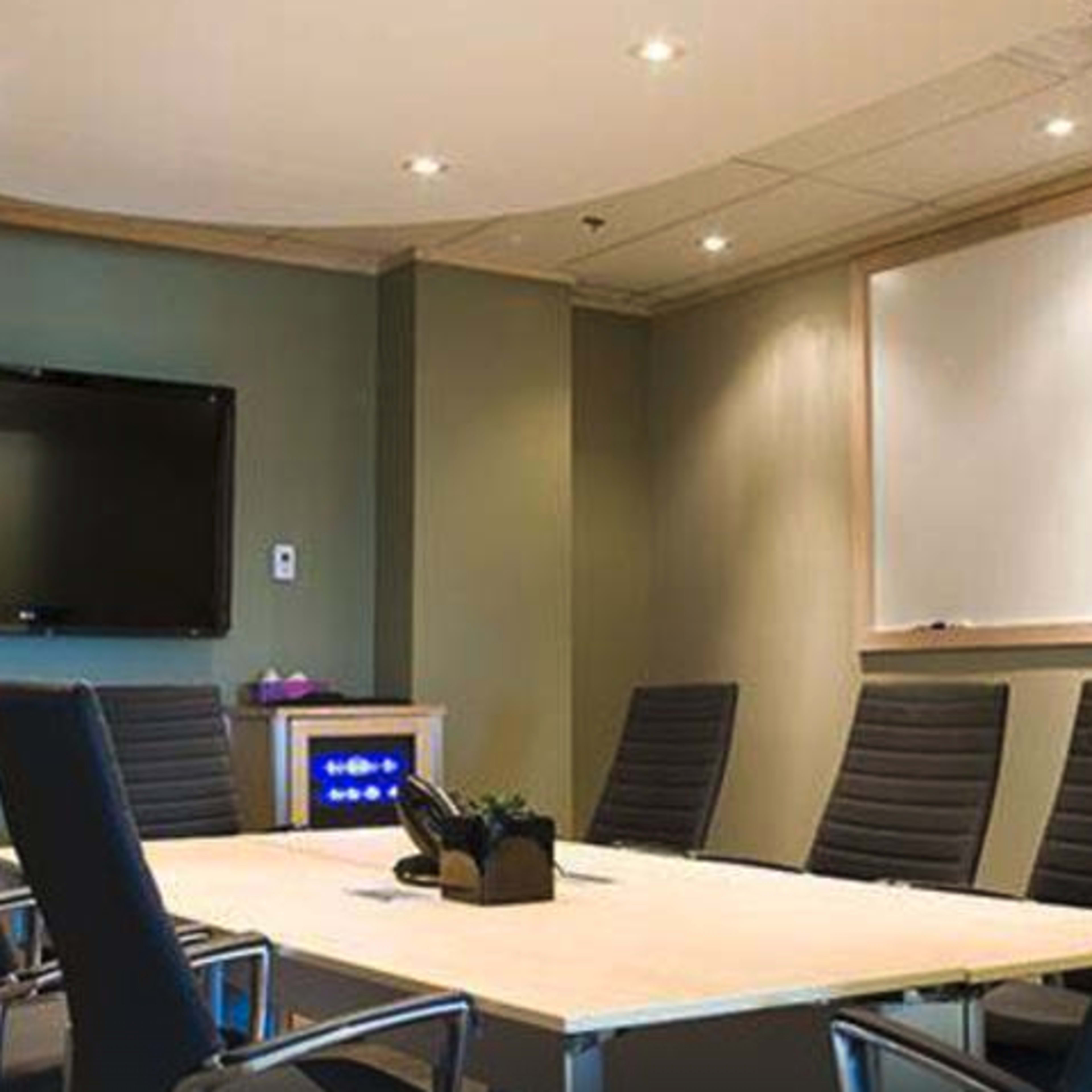 A modern conference room features a large table surrounded by black chairs, with a wall-mounted TV and a whiteboard in the background.