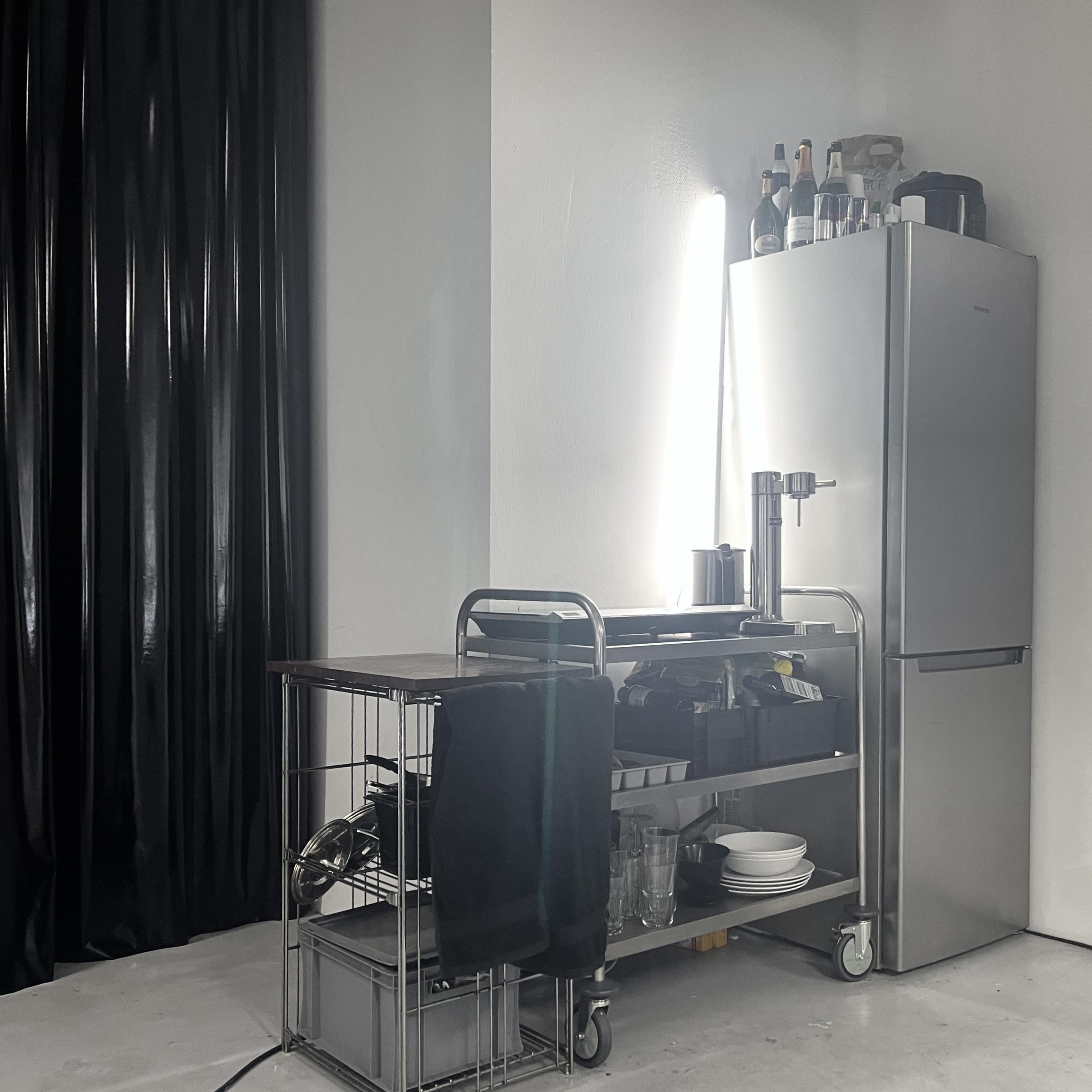A modern kitchen with a stainless steel refrigerator, a kitchen cart with various utensils, and a window covered by black curtains.