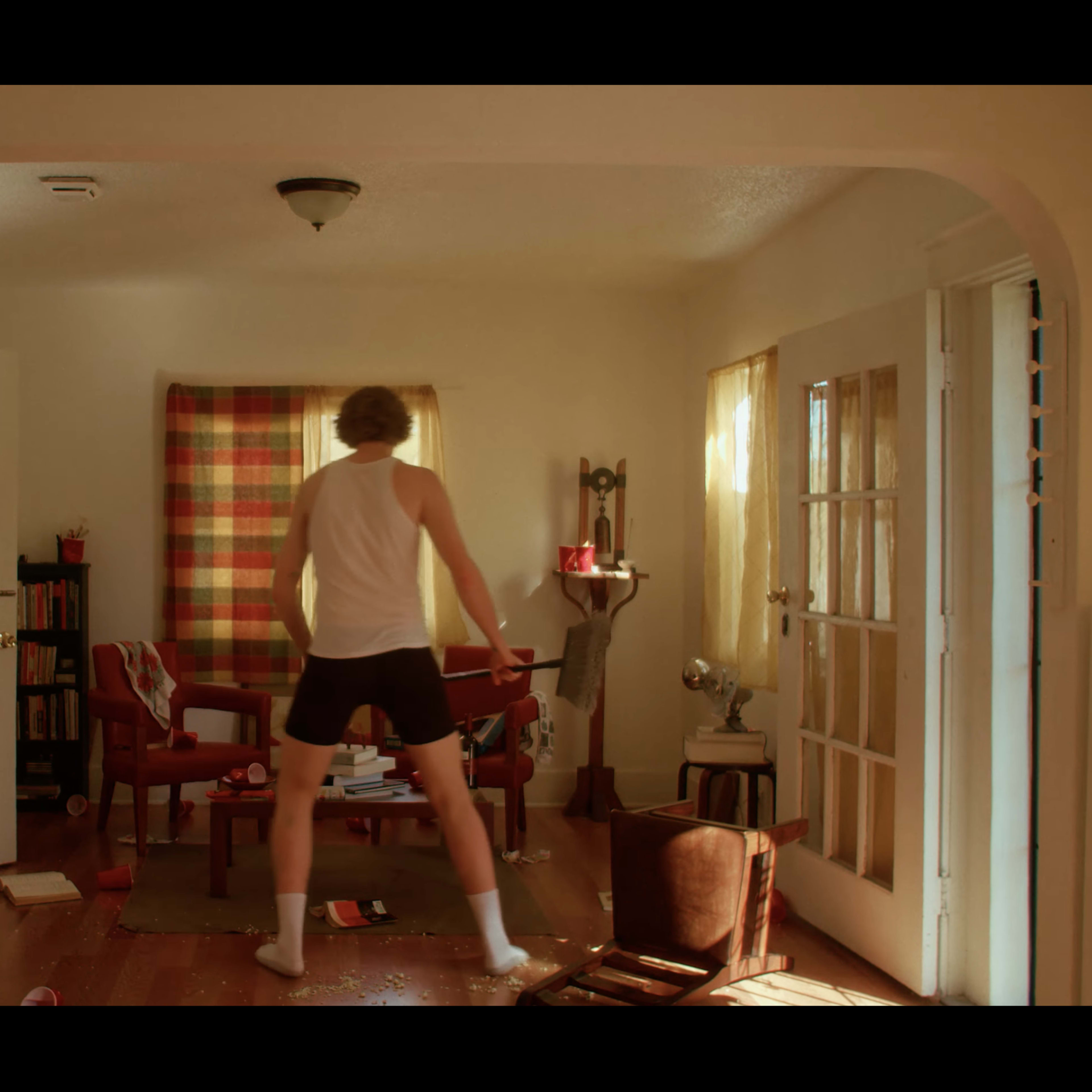 A man in a white tank top and shorts stands in a disordered living room with scattered books and furniture.