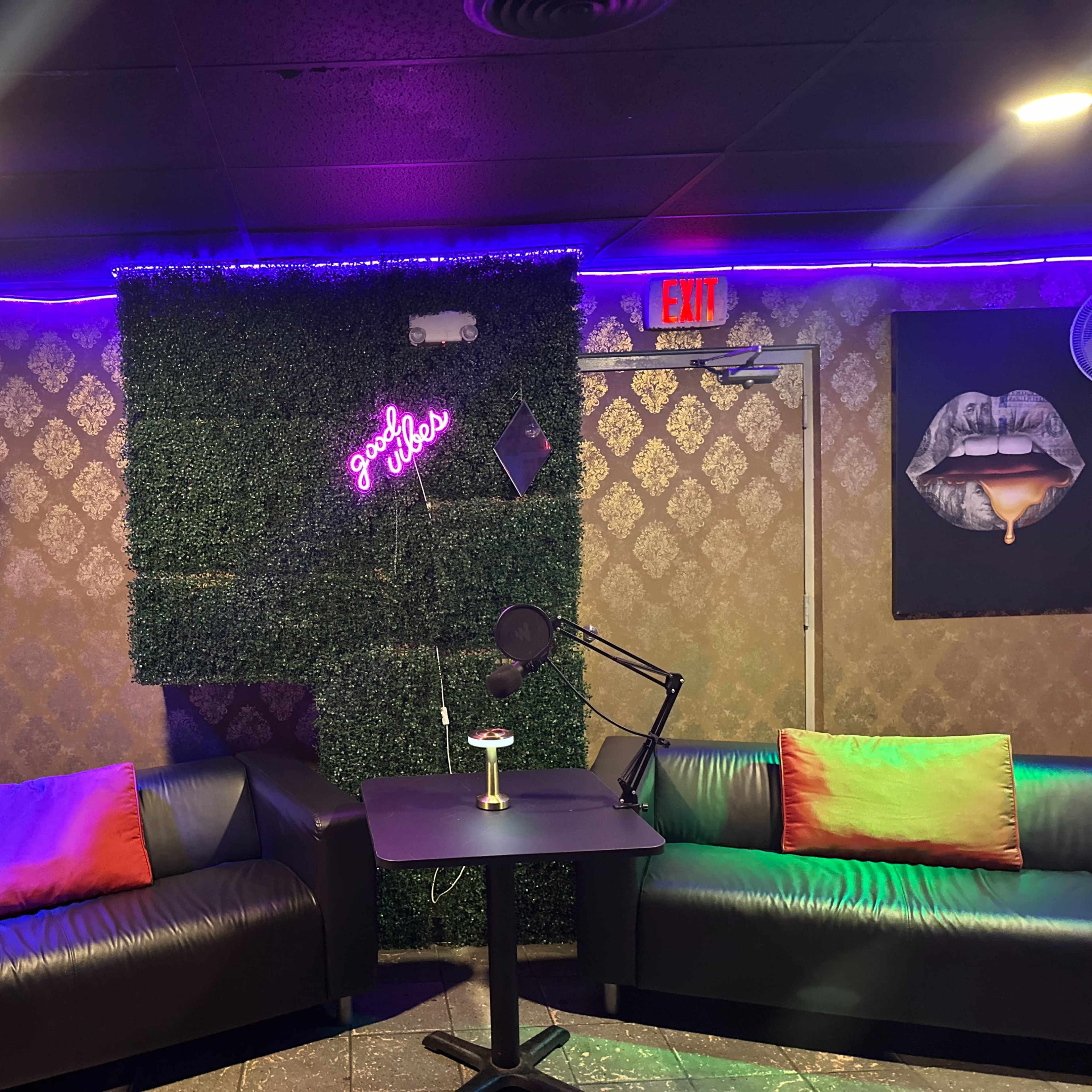 A small podcast recording studio featuring a green wall with lights, two black couches with colored pillows, and a microphone on a table.