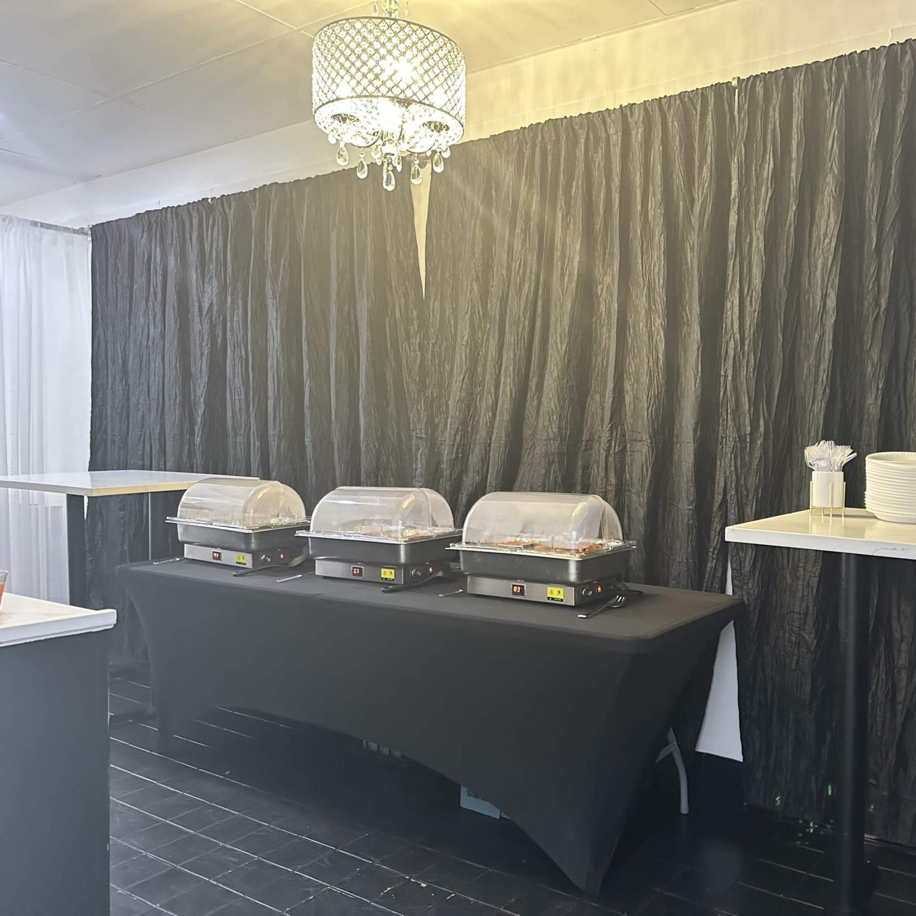 A buffet setup featuring three warming trays on a black tablecloth, with additional plates and utensils on a nearby table against a black backdrop.