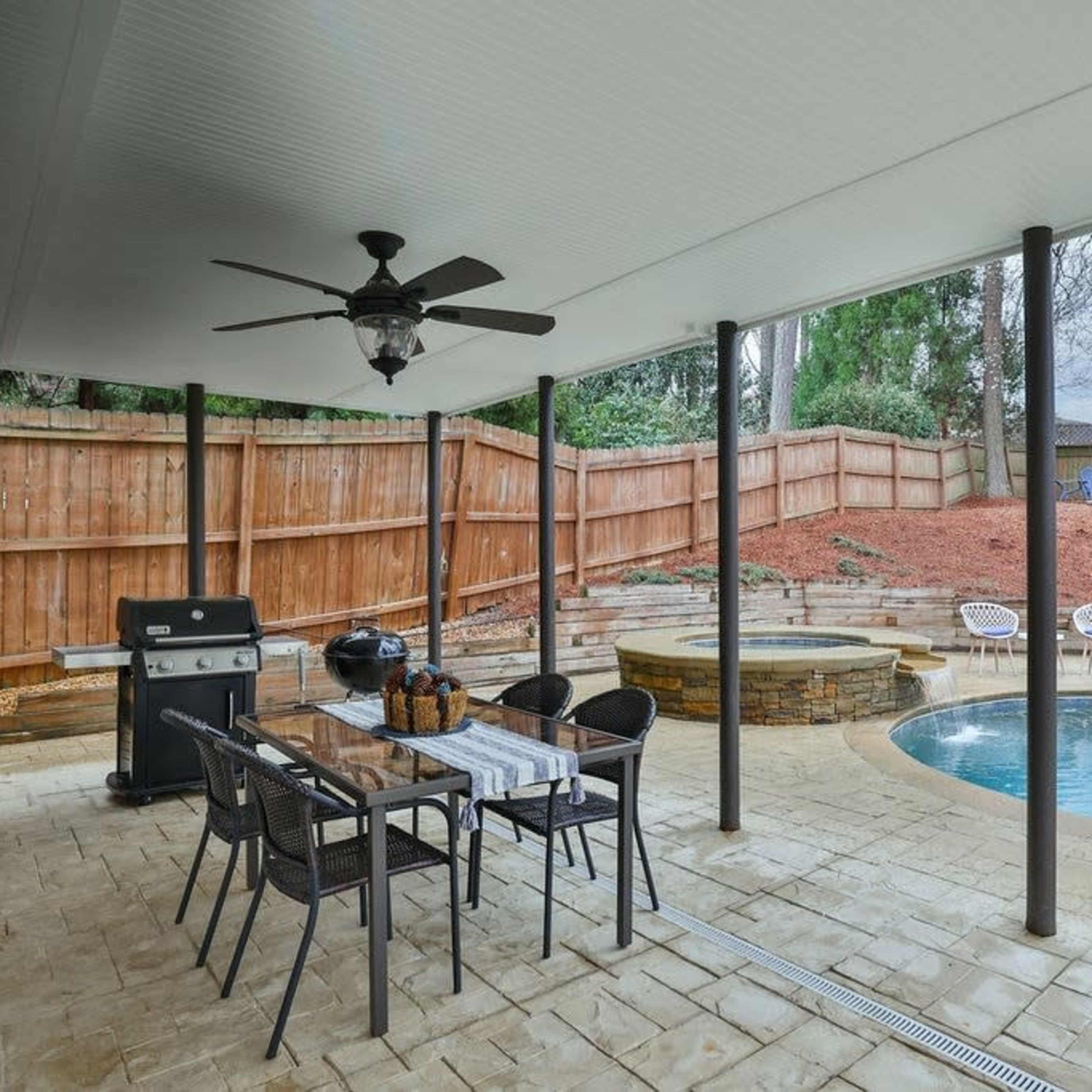 A covered patio area with a dining table, a grill, and a swimming pool featuring a spa, surrounded by a wooden fence and landscaped yard.
