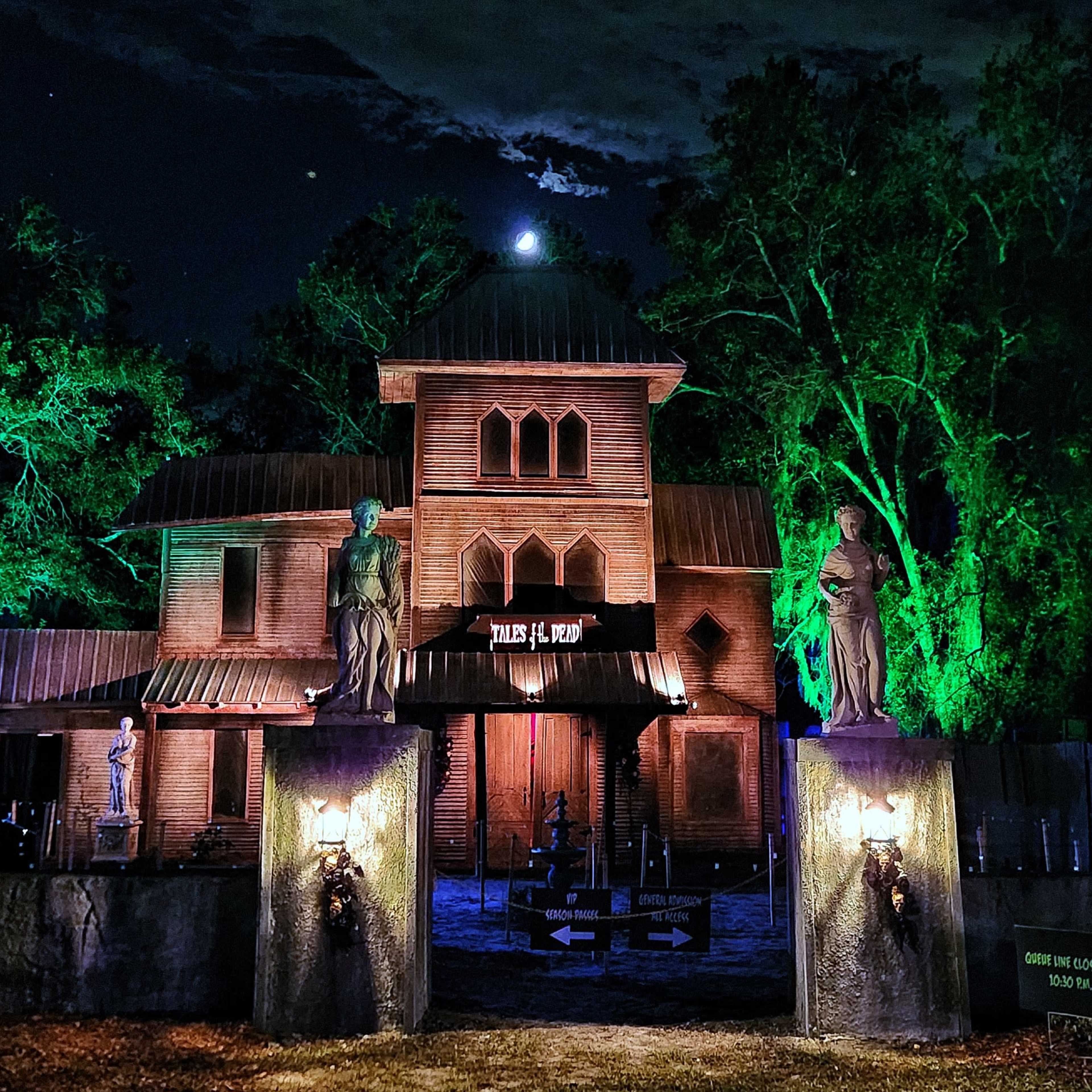 A haunted house attraction is illuminated with green lights at night, featuring statues at the entrance and a full moon in the background.