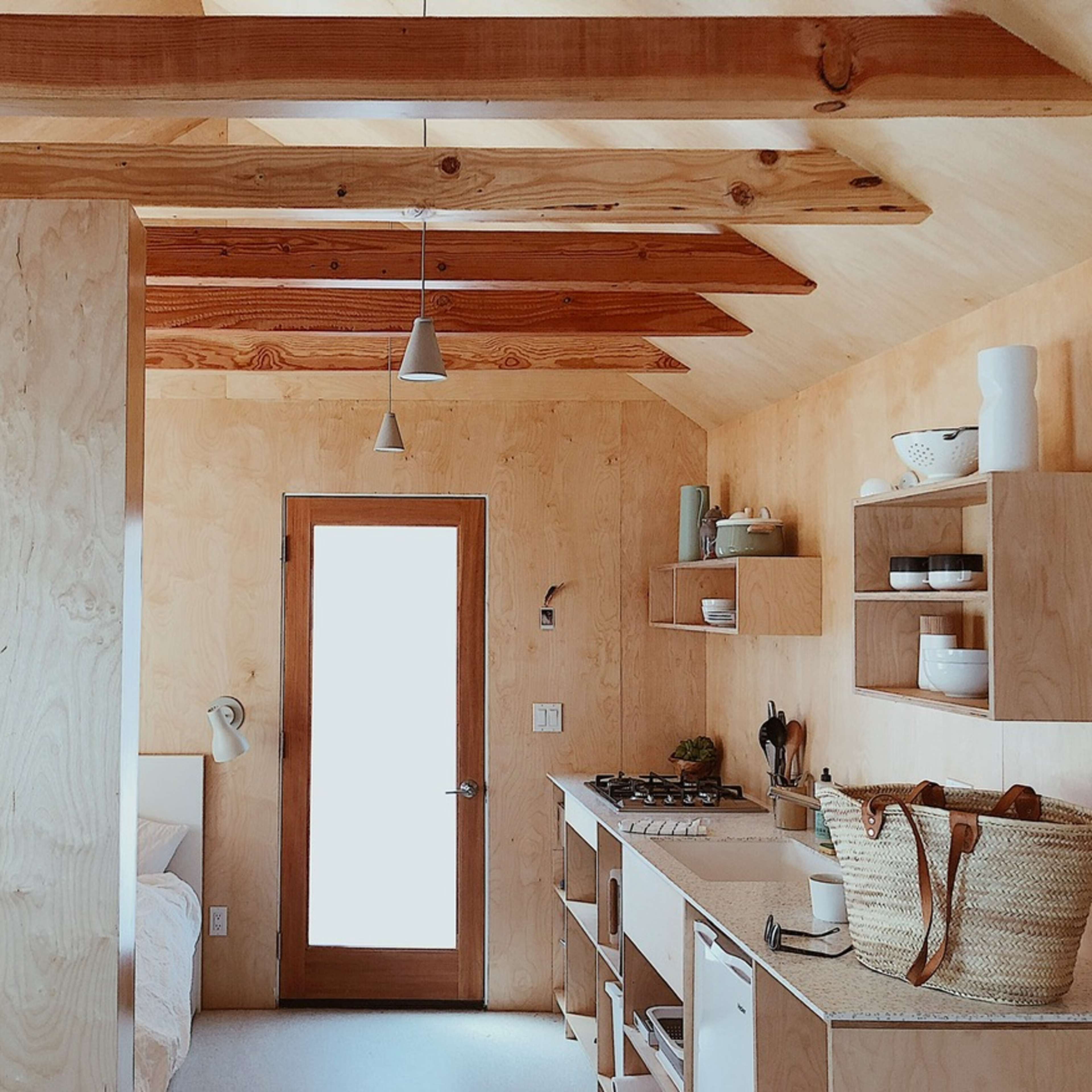 A minimalist interior of a small cabin kitchen with wooden beams, a doorway, and open shelving filled with dishware.