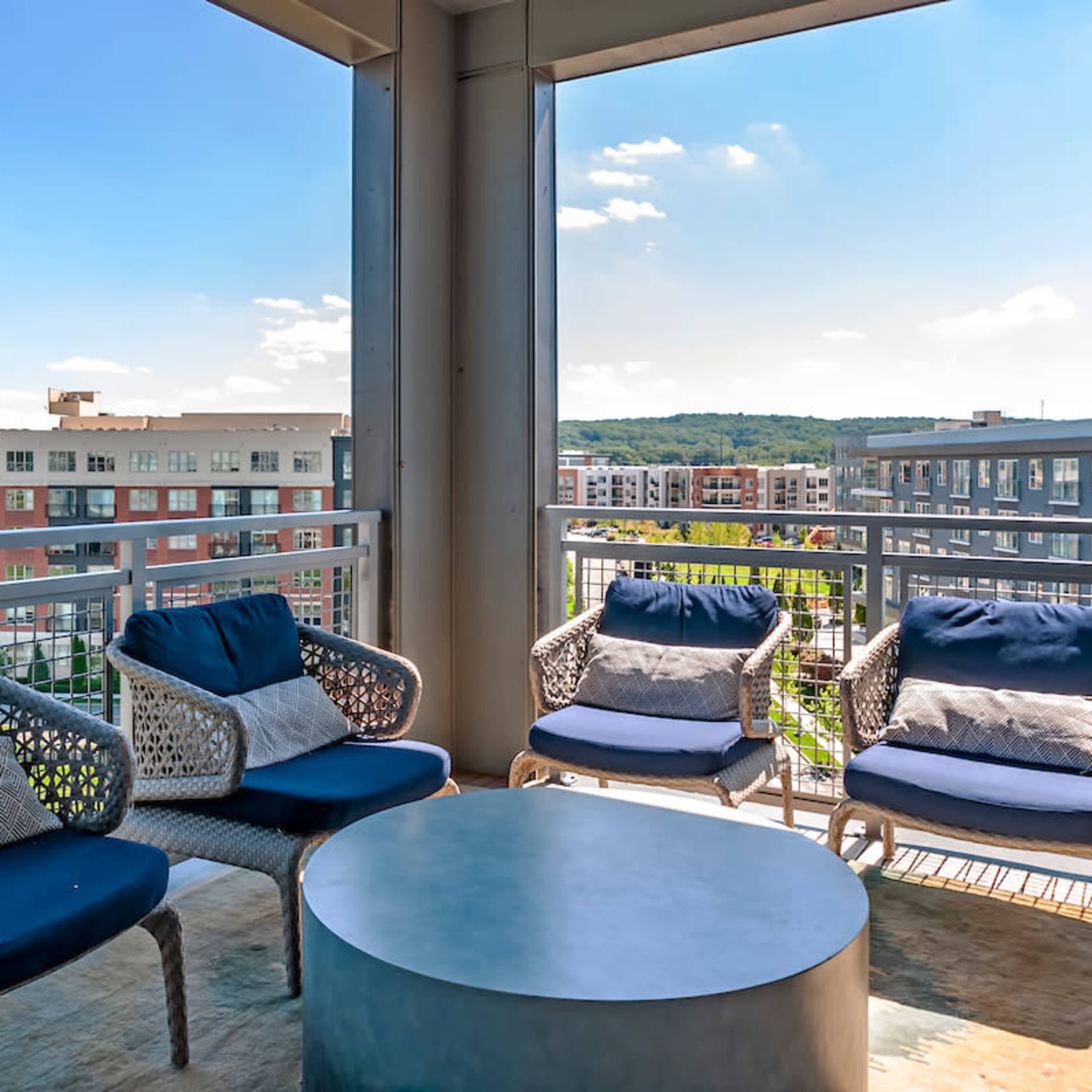 A balcony features four wicker chairs and a round table, overlooking an apartment complex and green hills in the distance.