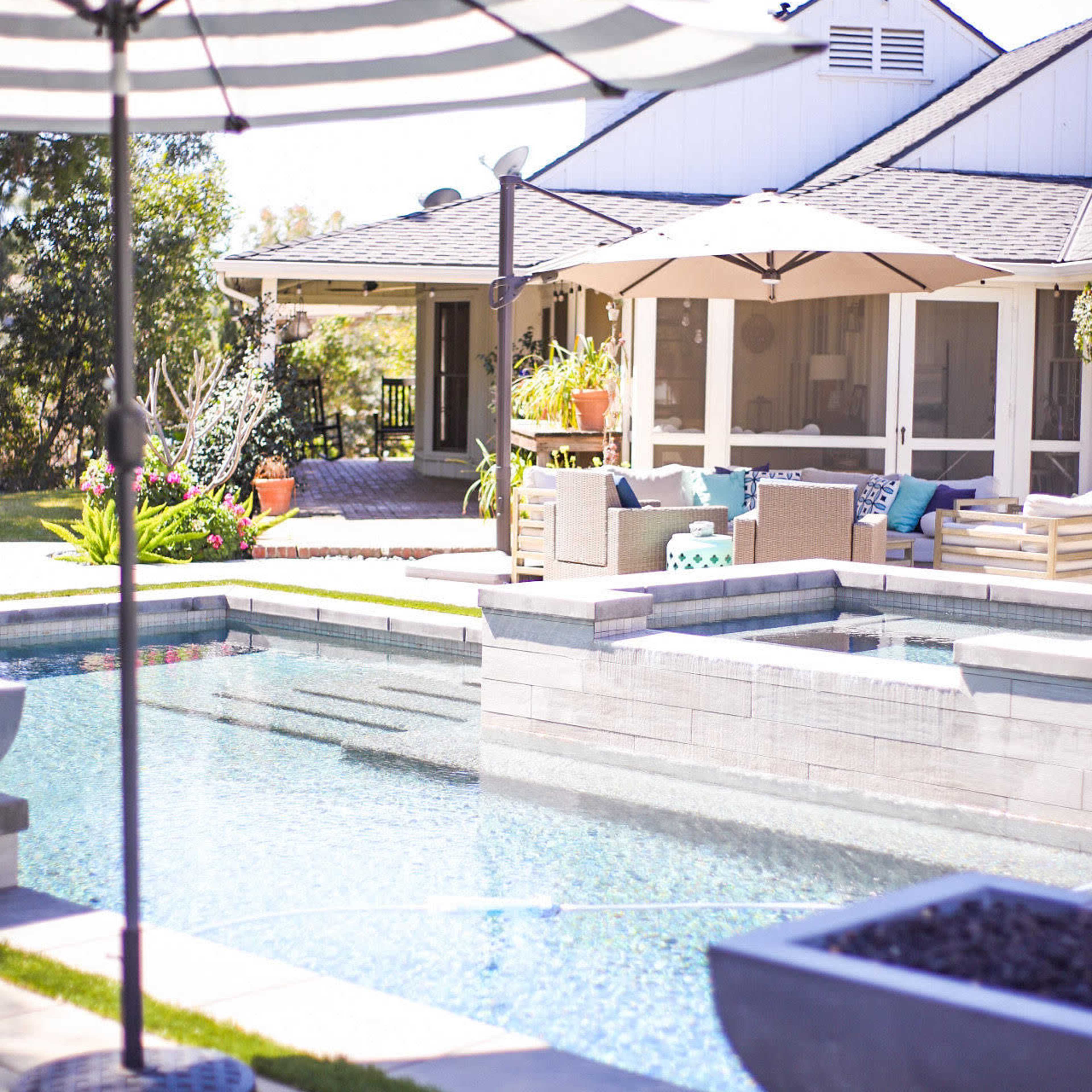 A backyard pool area with a striped umbrella, lounge furniture, and a house in the background surrounded by greenery.