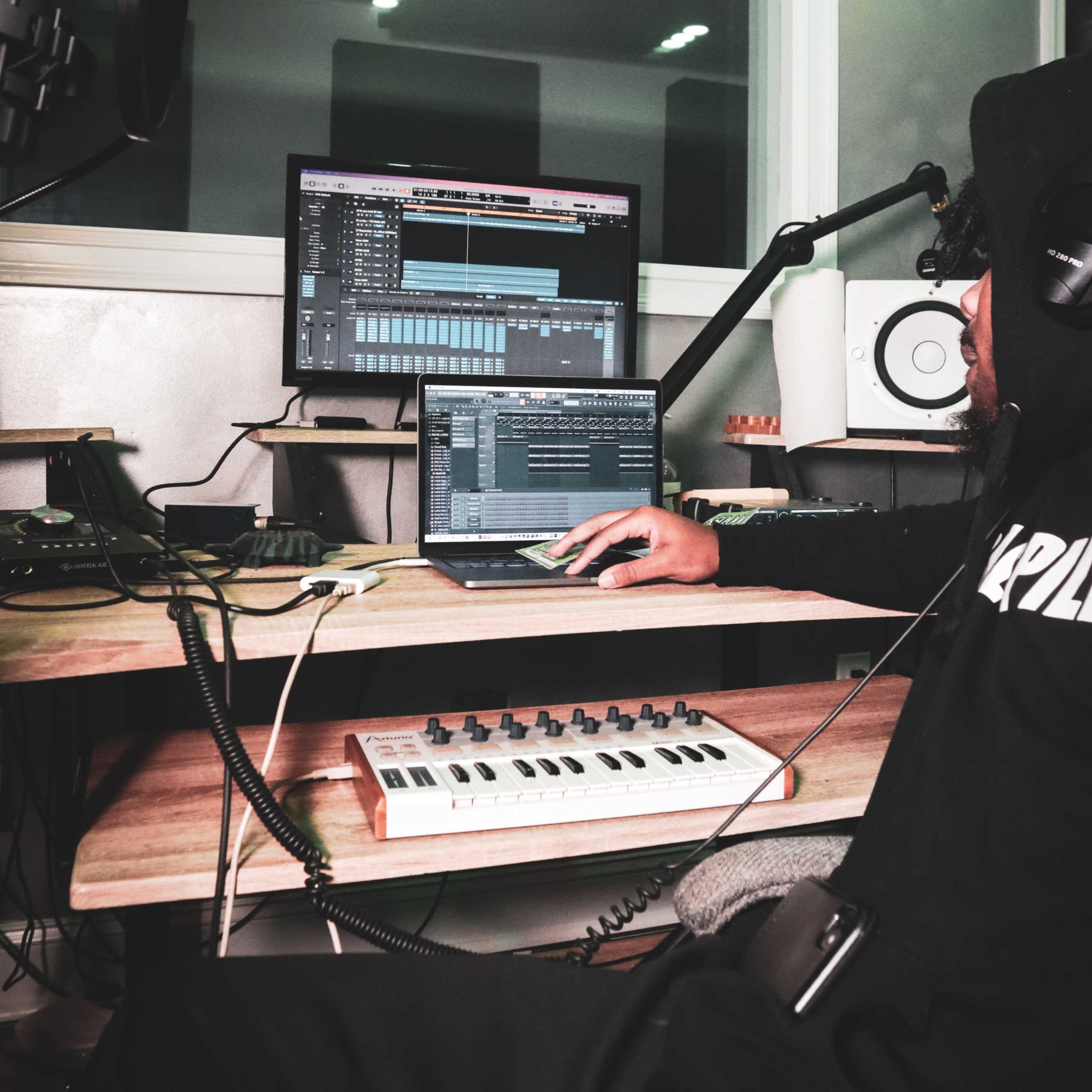 A person is sitting at a desk in a music studio, using a laptop and a MIDI keyboard while working on a digital audio workstation.