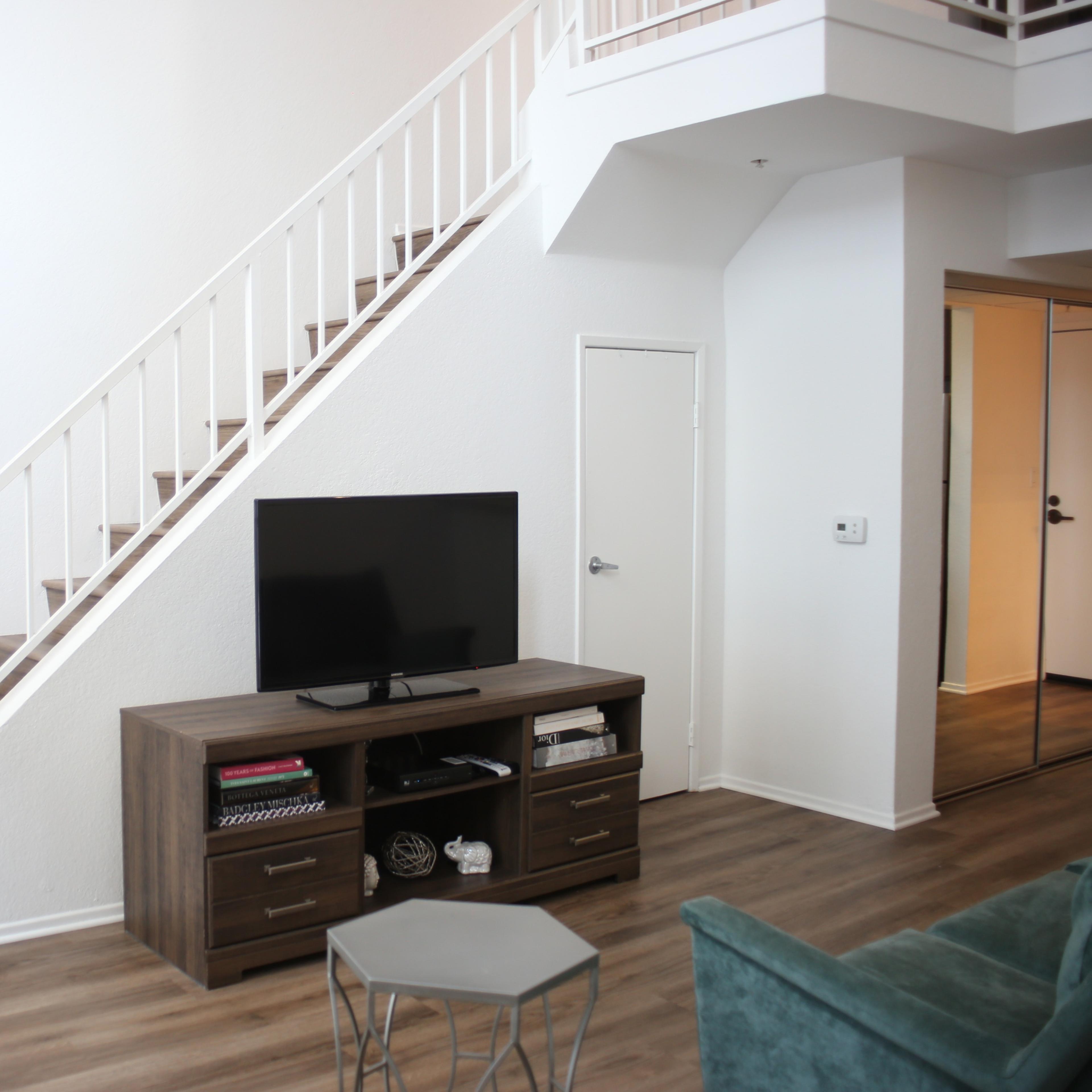 A living room with a television stand and a flat-screen TV positioned against a wall near a staircase leading to an upper level.