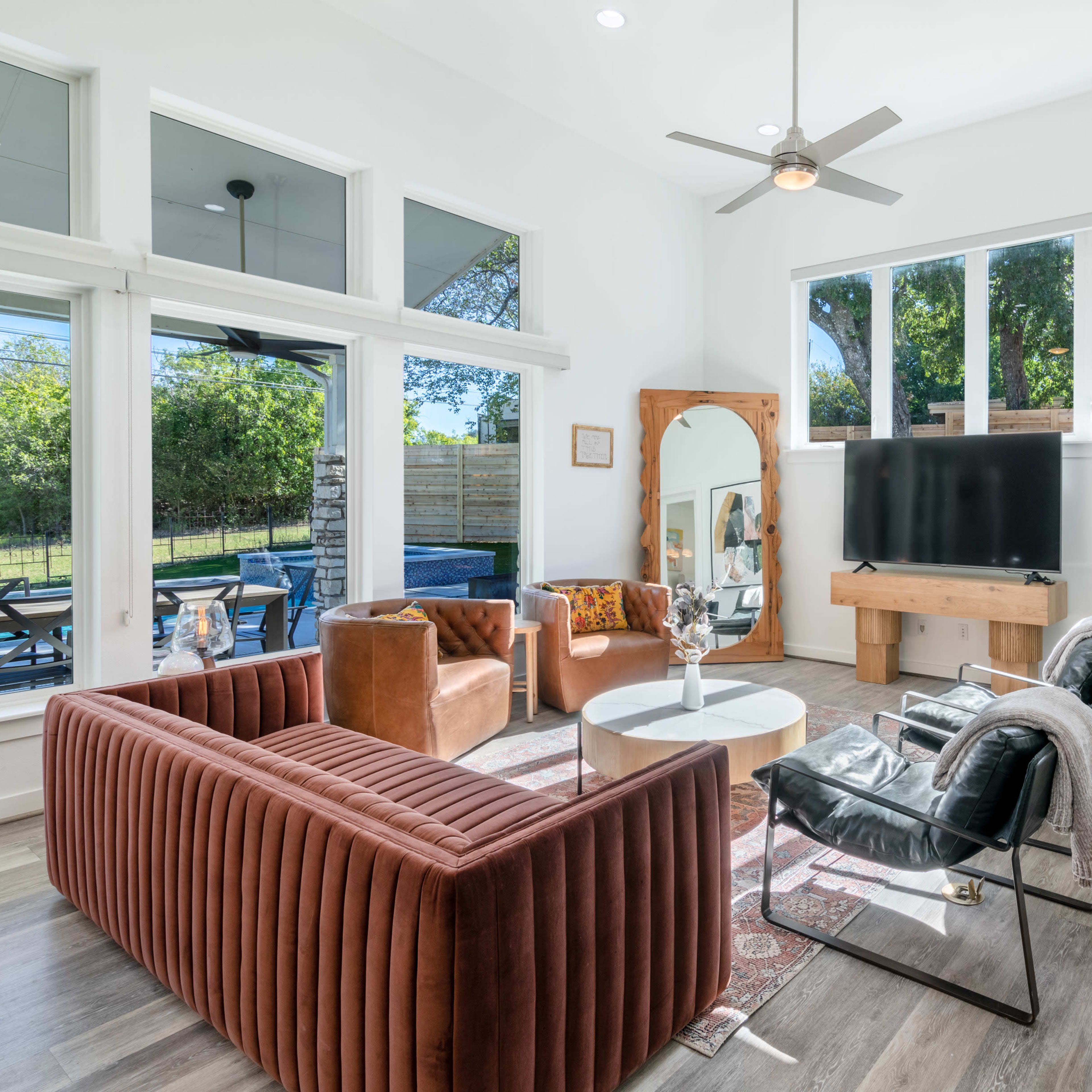 The living room features a modern design with a brown velvet sofa, leather armchairs, a large wall-mounted TV, and abundant natural light from large windows.