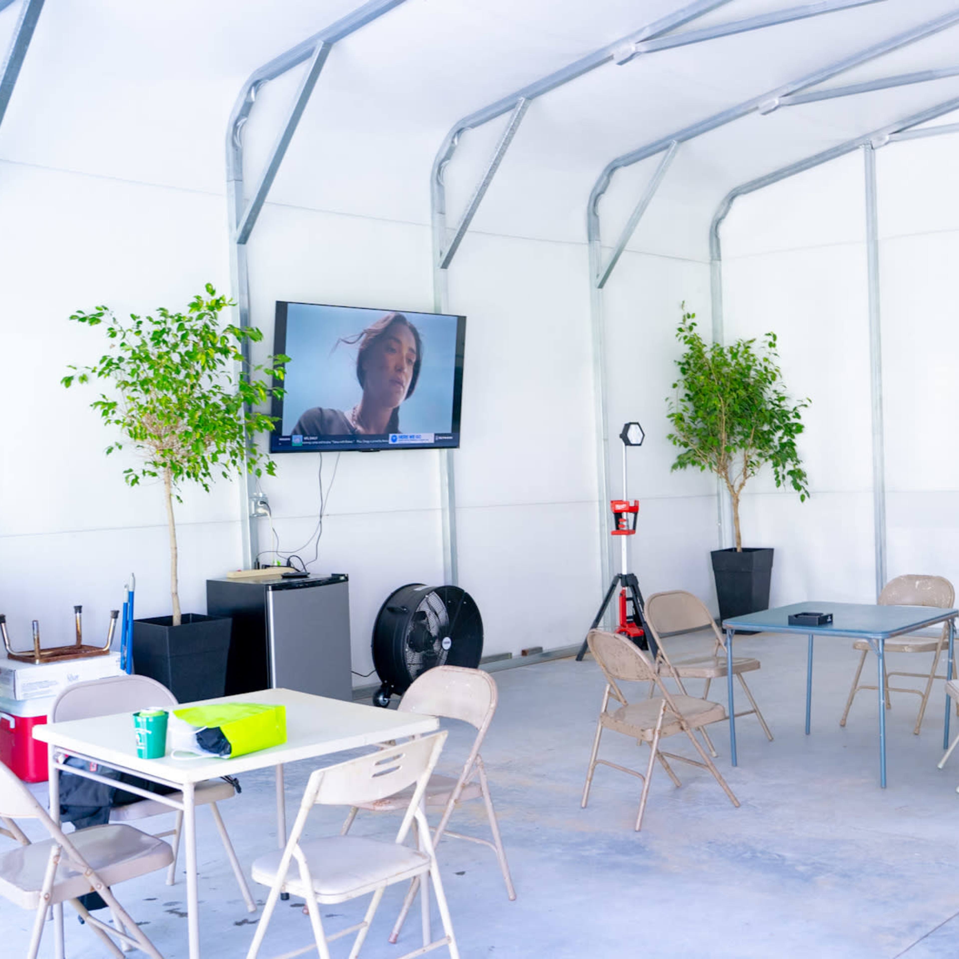 An indoor gathering space features metal chairs and tables, potted plants, a television mounted on the wall, and a fan.