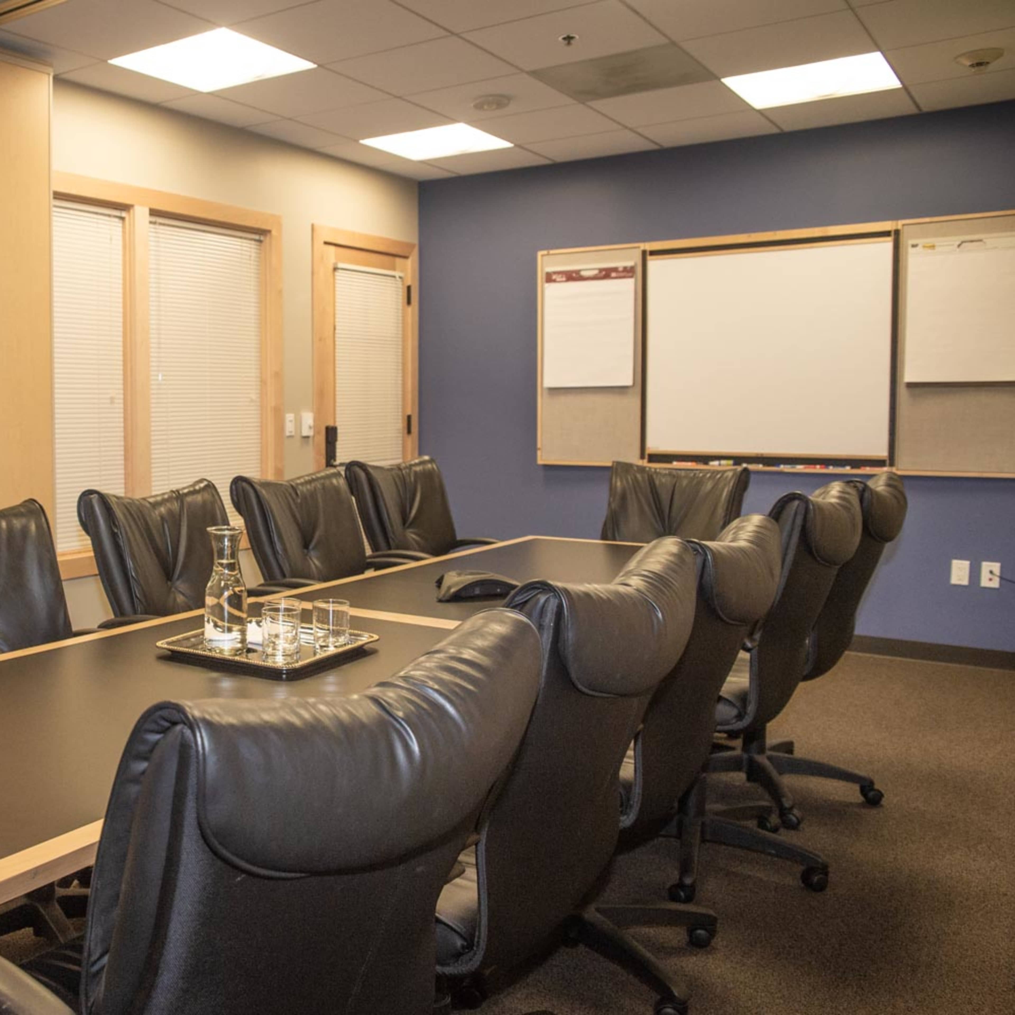 A conference room with a large wooden table surrounded by black leather chairs, featuring a whiteboard and presentation boards on the walls.