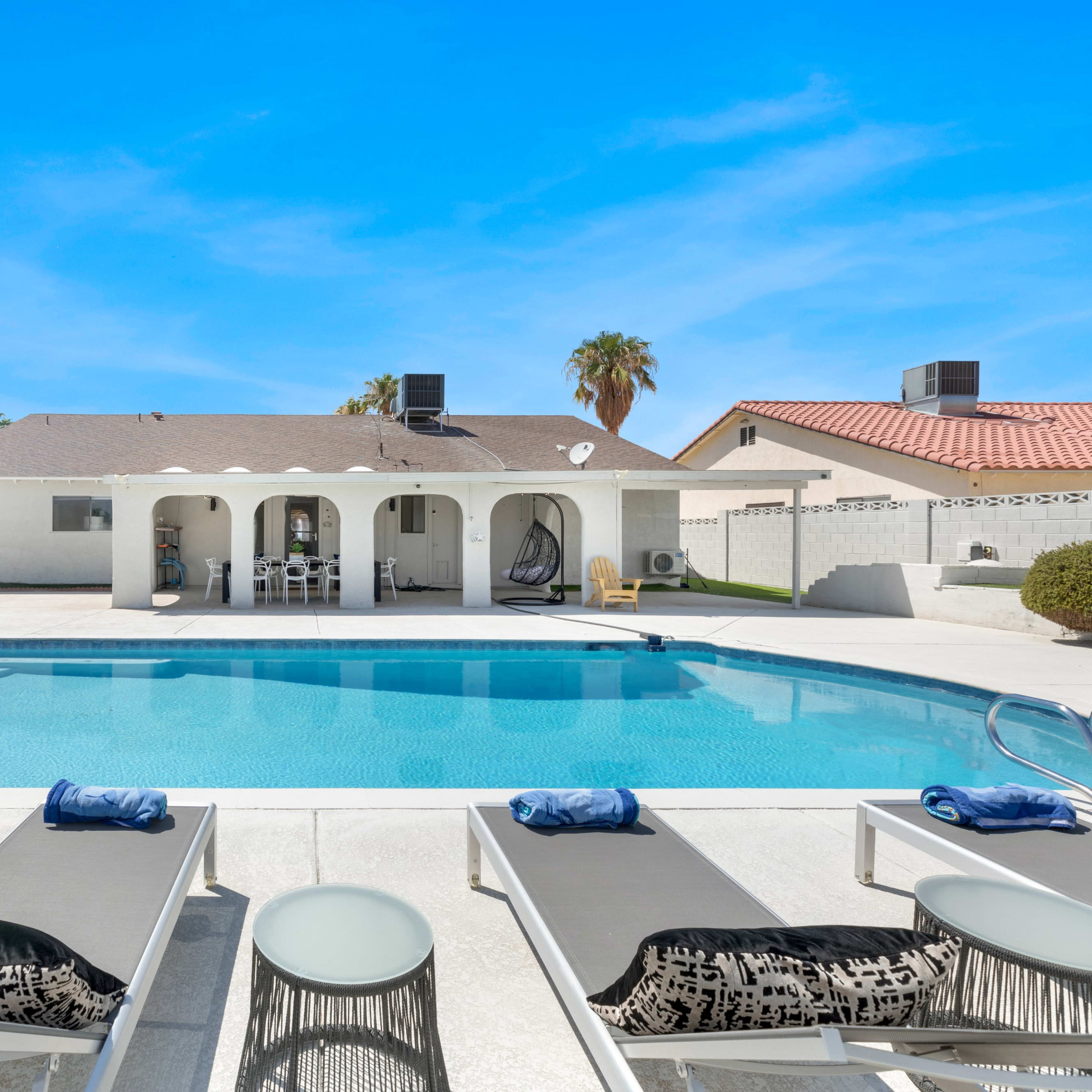 A clear blue swimming pool is flanked by lounge chairs and features a single-story home with arched openings in the background.