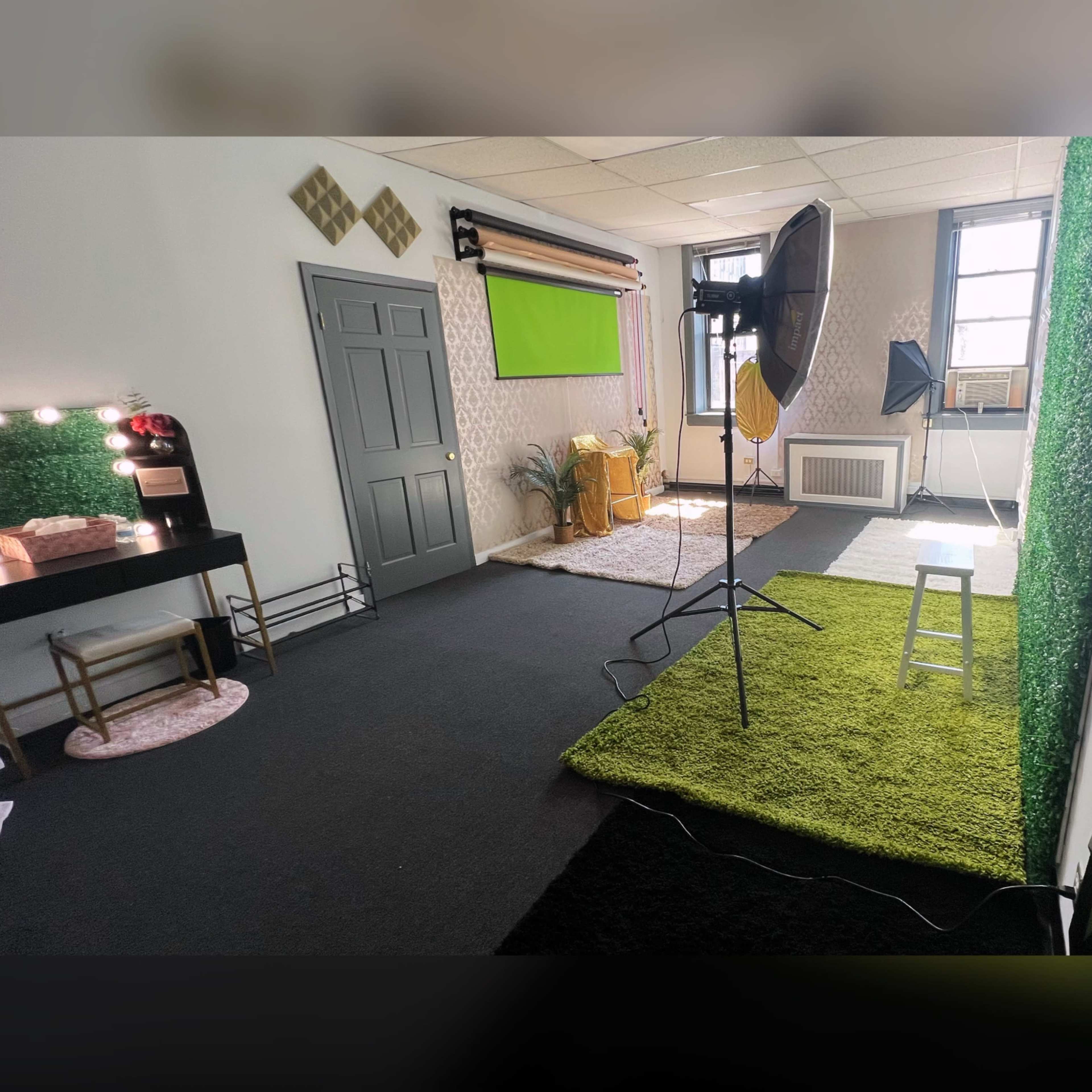 A studio space featuring a green screen setup, a large softbox light, a chair, and decorative elements, with a blend of grass-like flooring and a dark carpet.