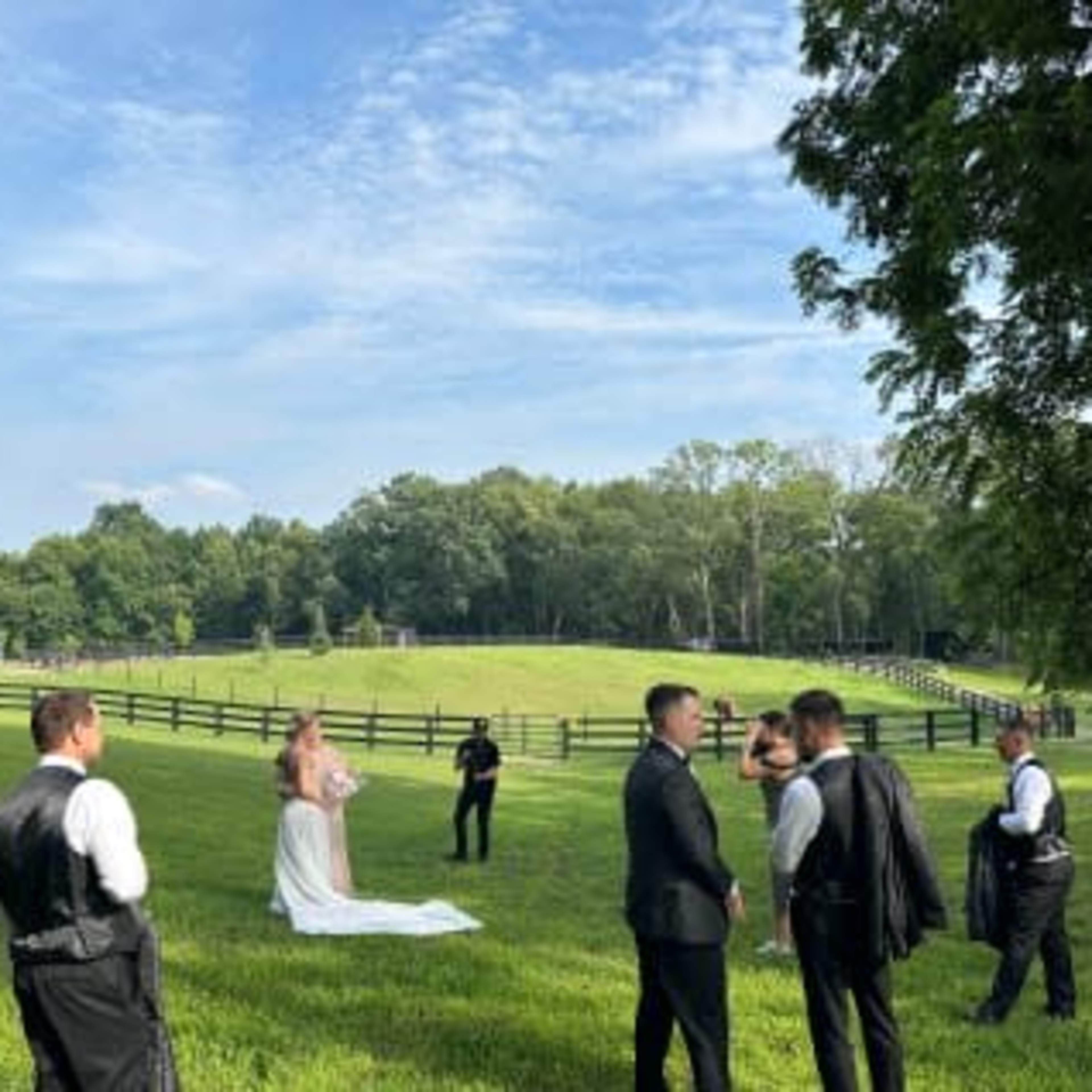 A bride in a white dress stands on a grassy field surrounded by several men dressed in formal attire, with trees and a fence in the background.