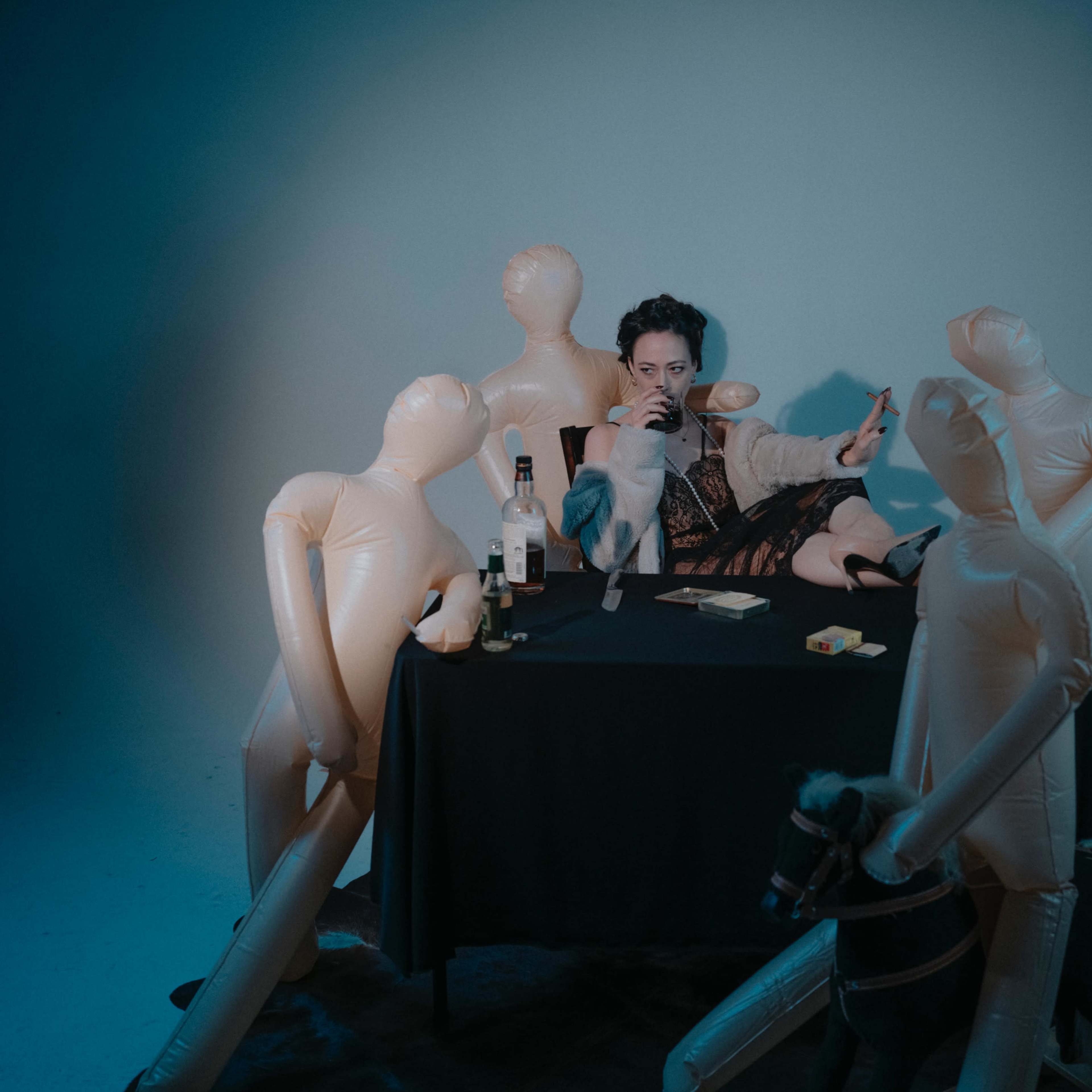 A person sits at a table surrounded by inflatable dolls, holding a drink with playful expressions on their faces.