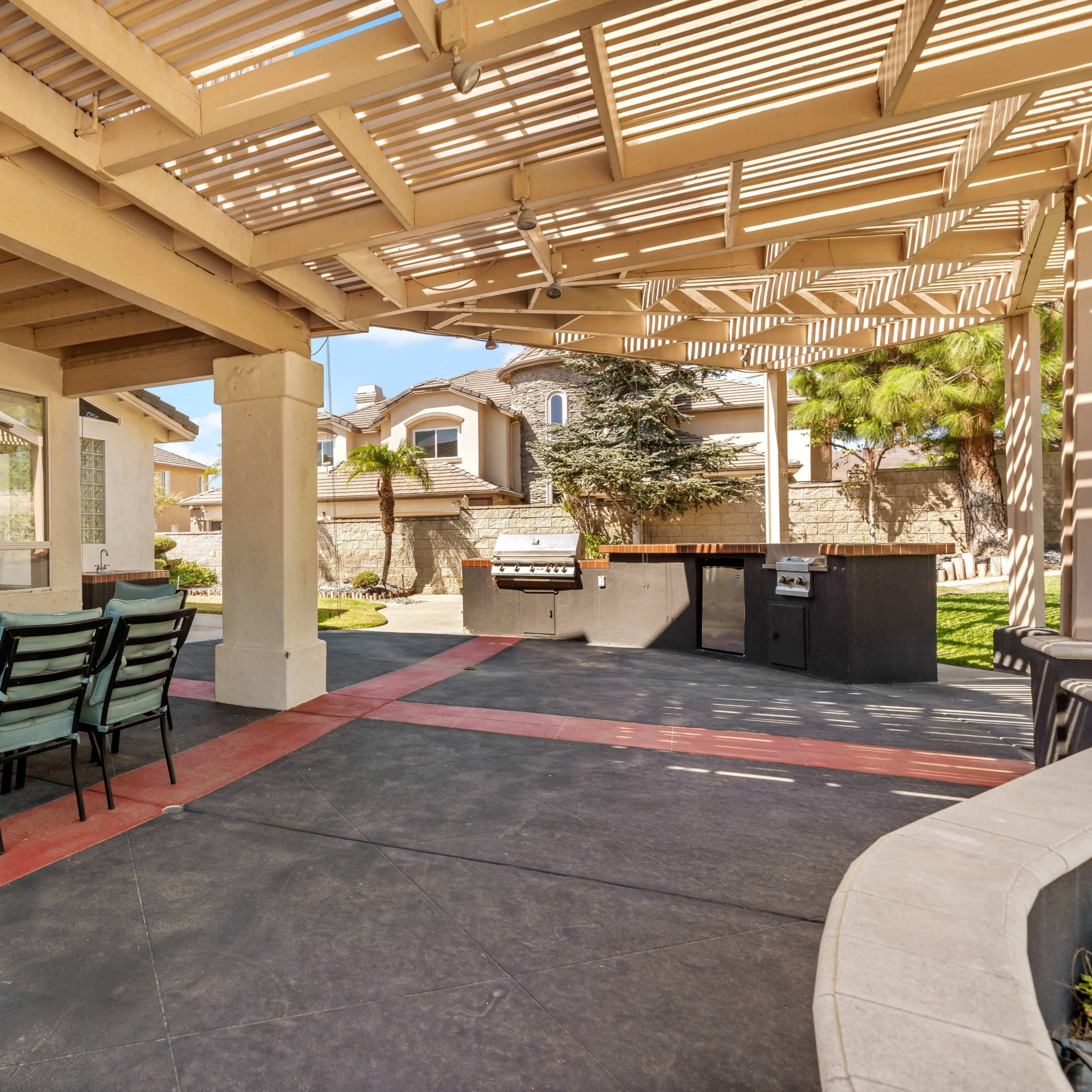 A covered patio area features a dining table with chairs and a barbecue station in a residential backyard.