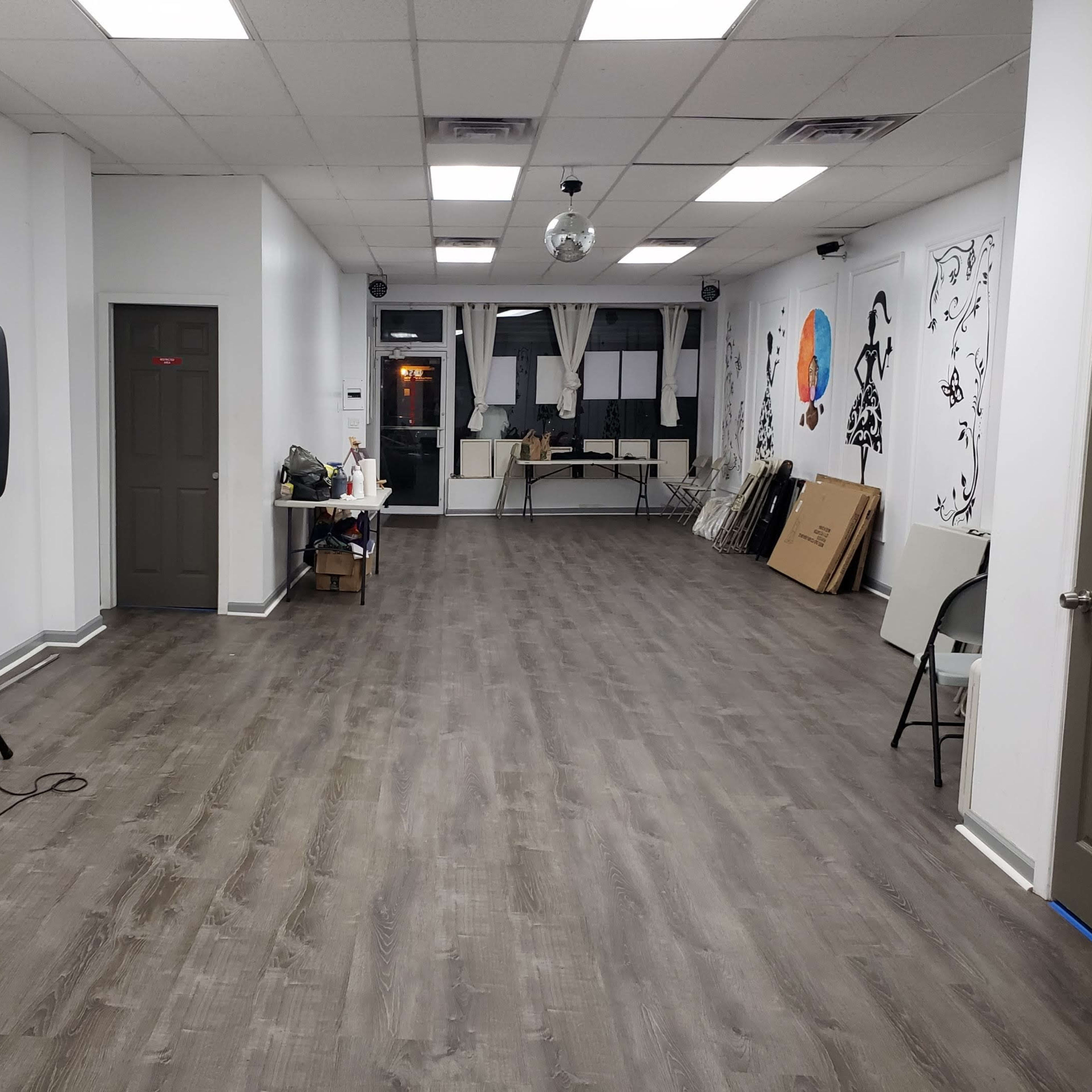 An empty interior space with wooden flooring, white walls, and a speaker on a stand near the entrance, along with various items stacked on tables.