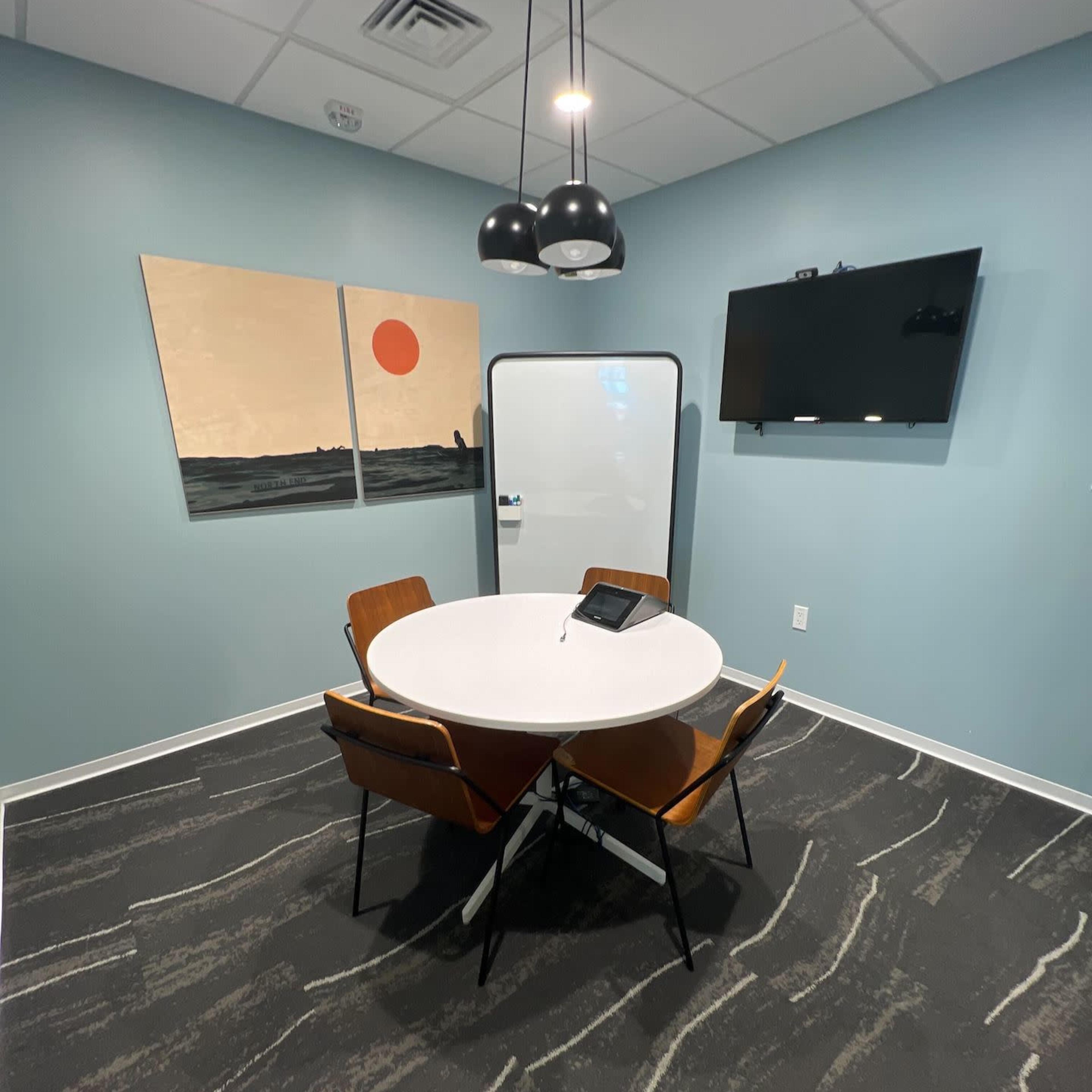 A small conference room features a round table, four chairs, a flat-screen TV, a whiteboard, and two abstract paintings on the wall.