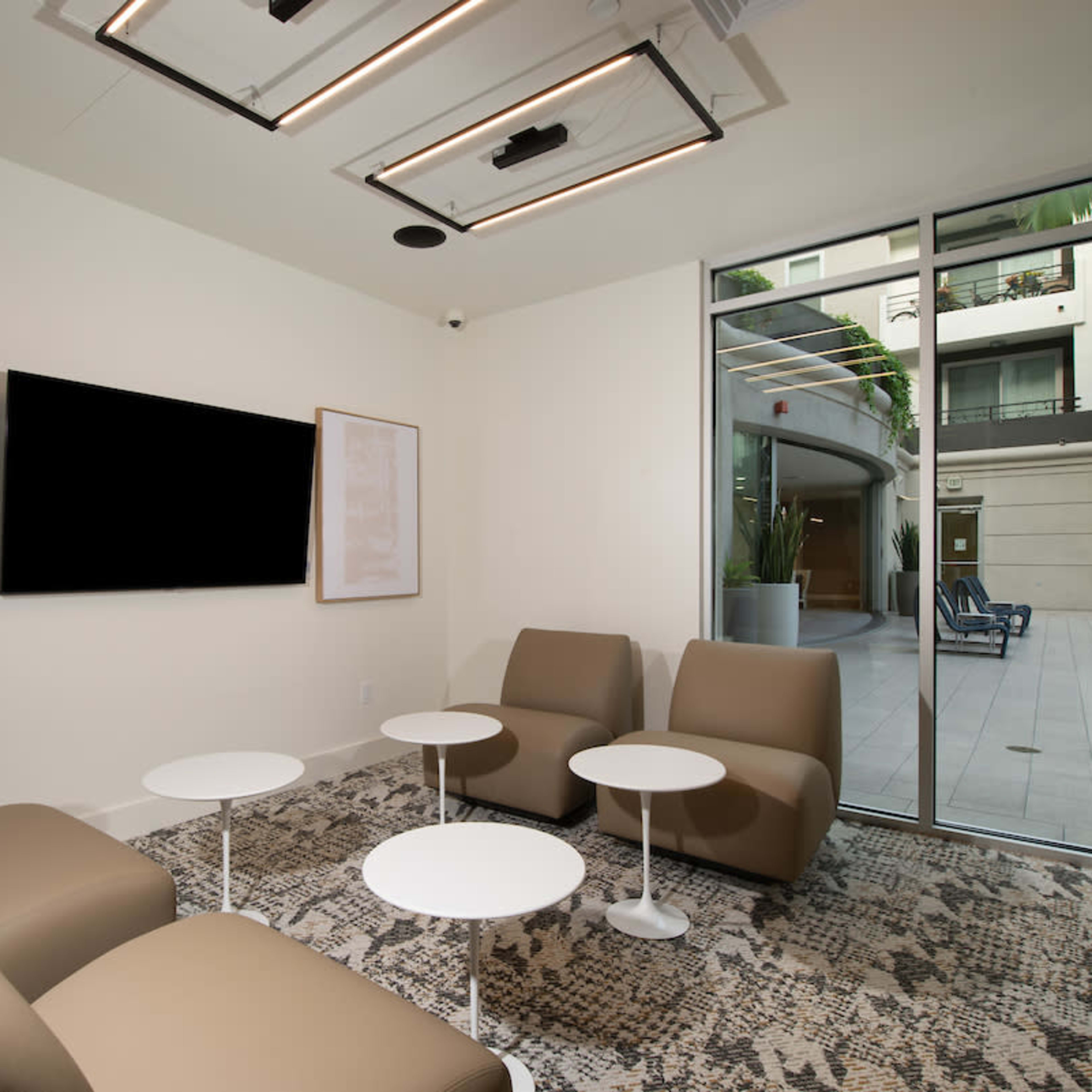 A modern lounge area featuring beige chairs, white tables, and a large window overlooking an exterior space.