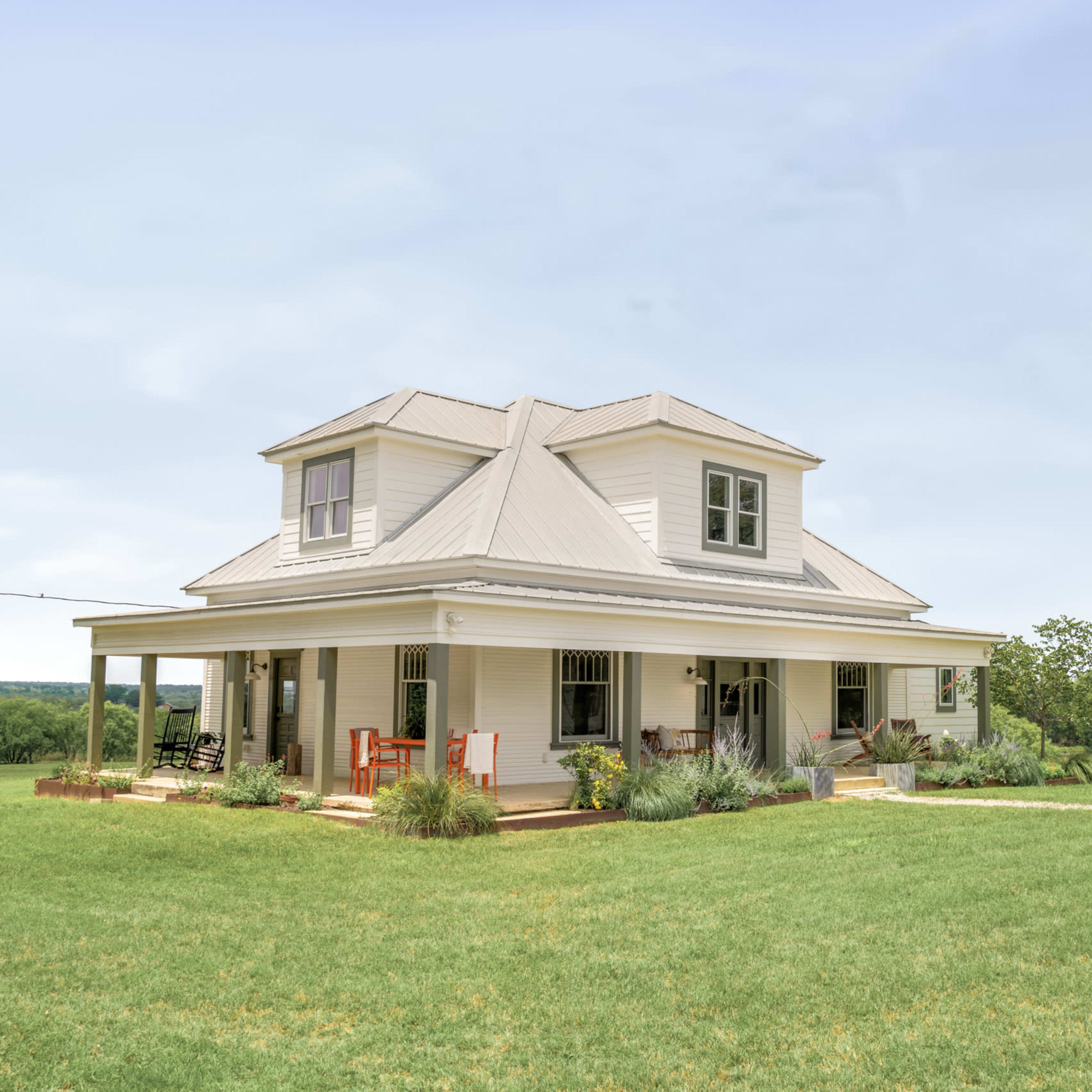 A white two-story house with a metal roof is surrounded by a green lawn and small trees in an open field.