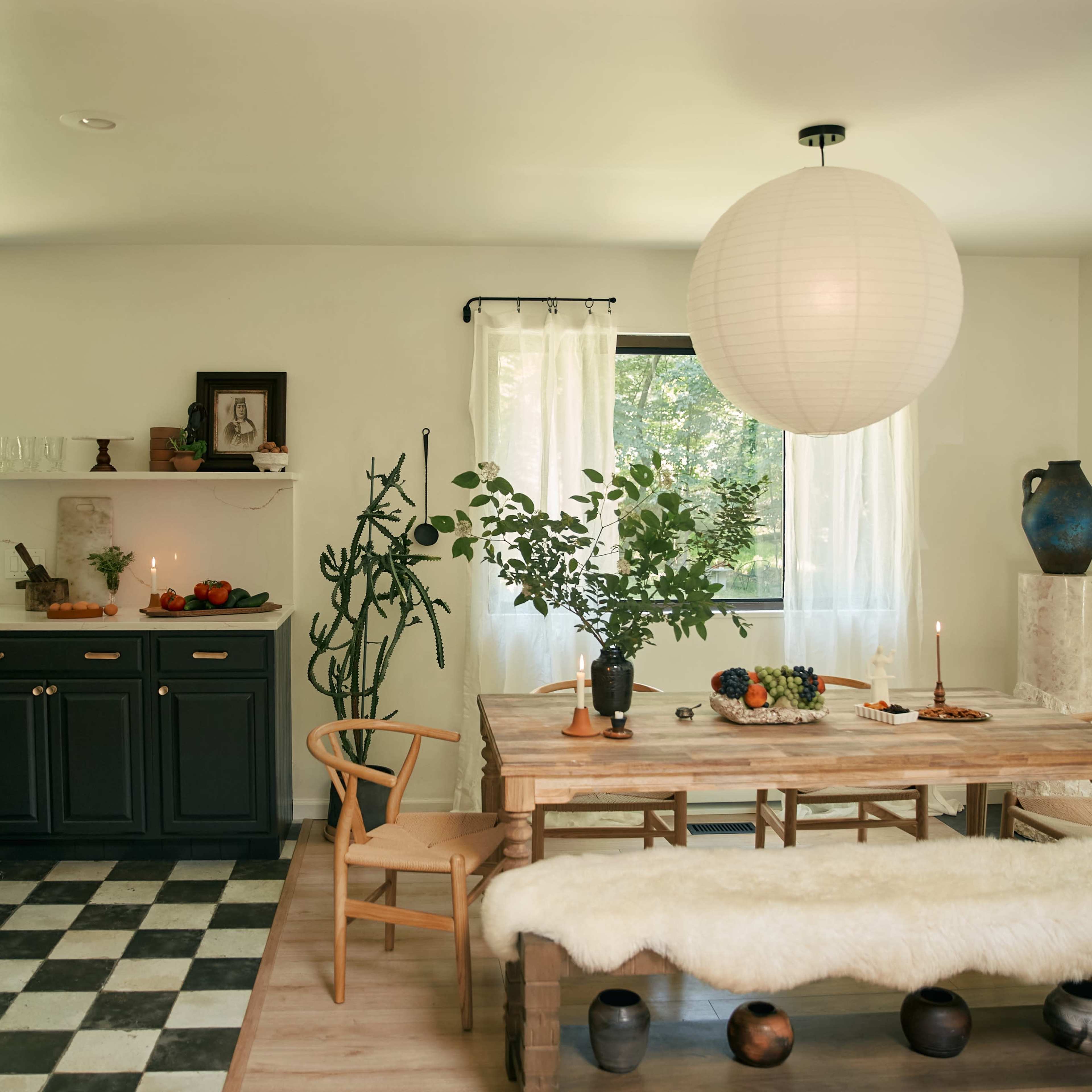 A modern dining area features a wooden table with a centerpiece, surrounded by chairs, and a kitchen in the background with dark cabinetry and a window revealing greenery outside.