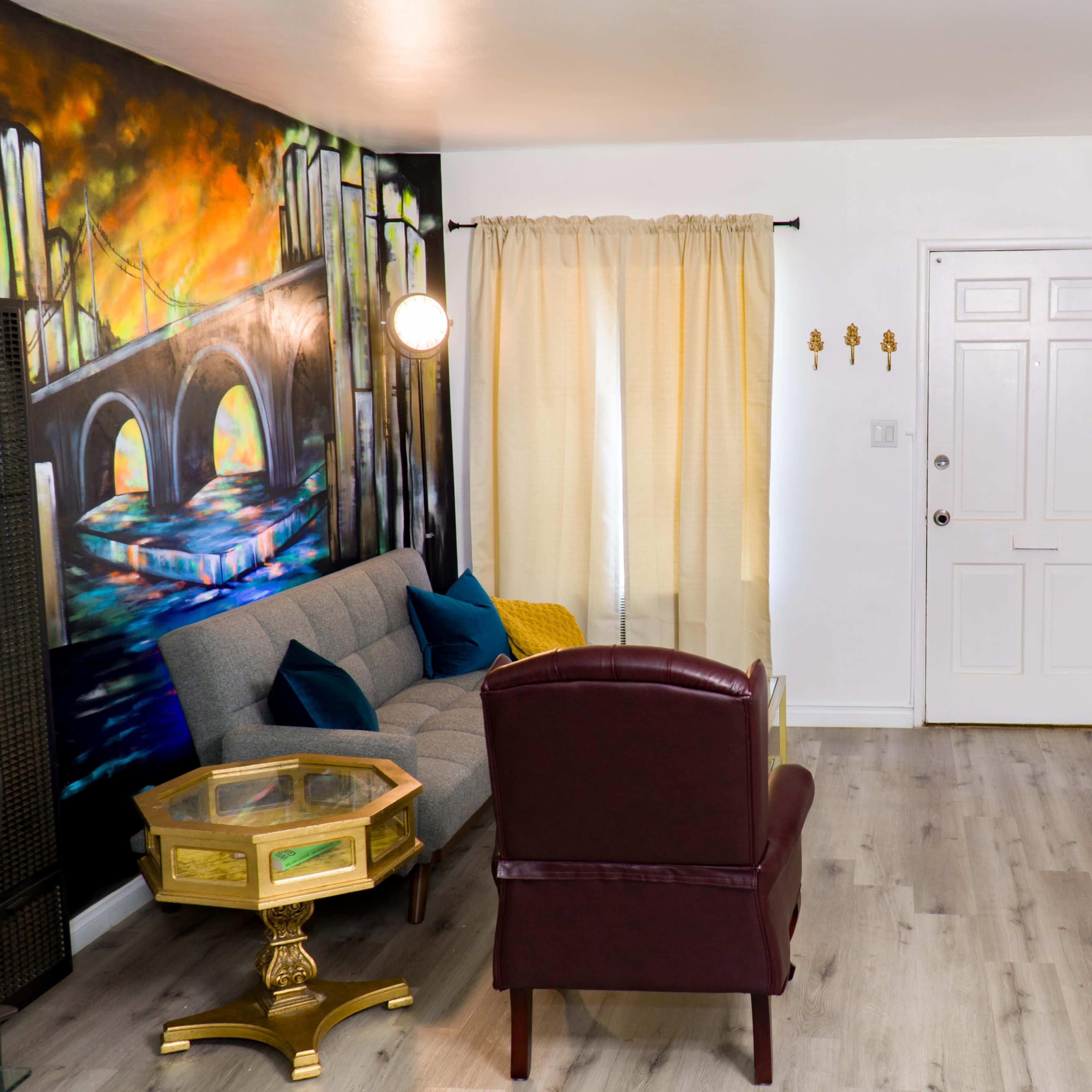 The room features a gray sofa, a burgundy armchair, and colorful mural artwork on two walls, with a door and a mounted television visible.