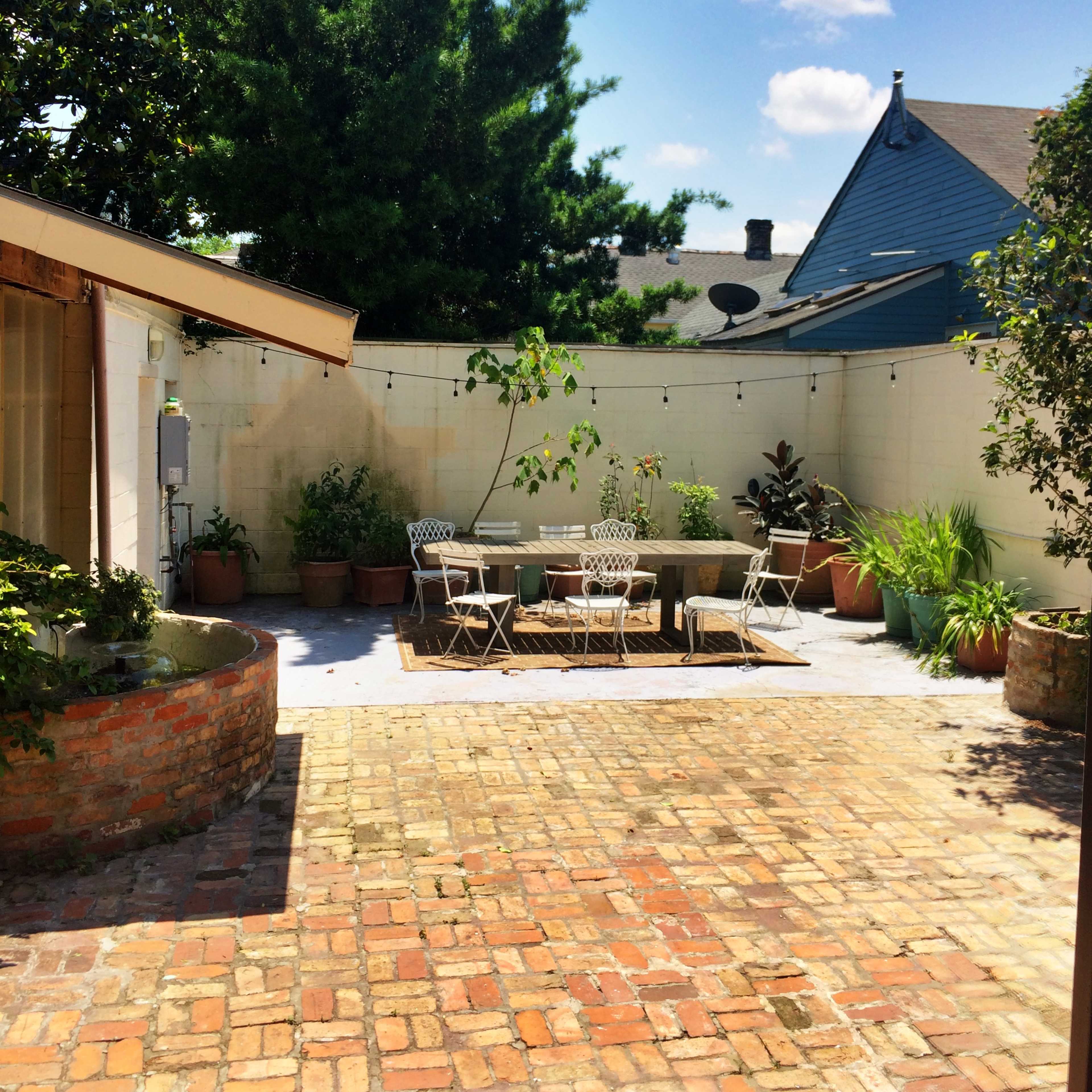 A brick-paved patio features a circular table and chairs surrounded by potted plants and a wall with a green vine.