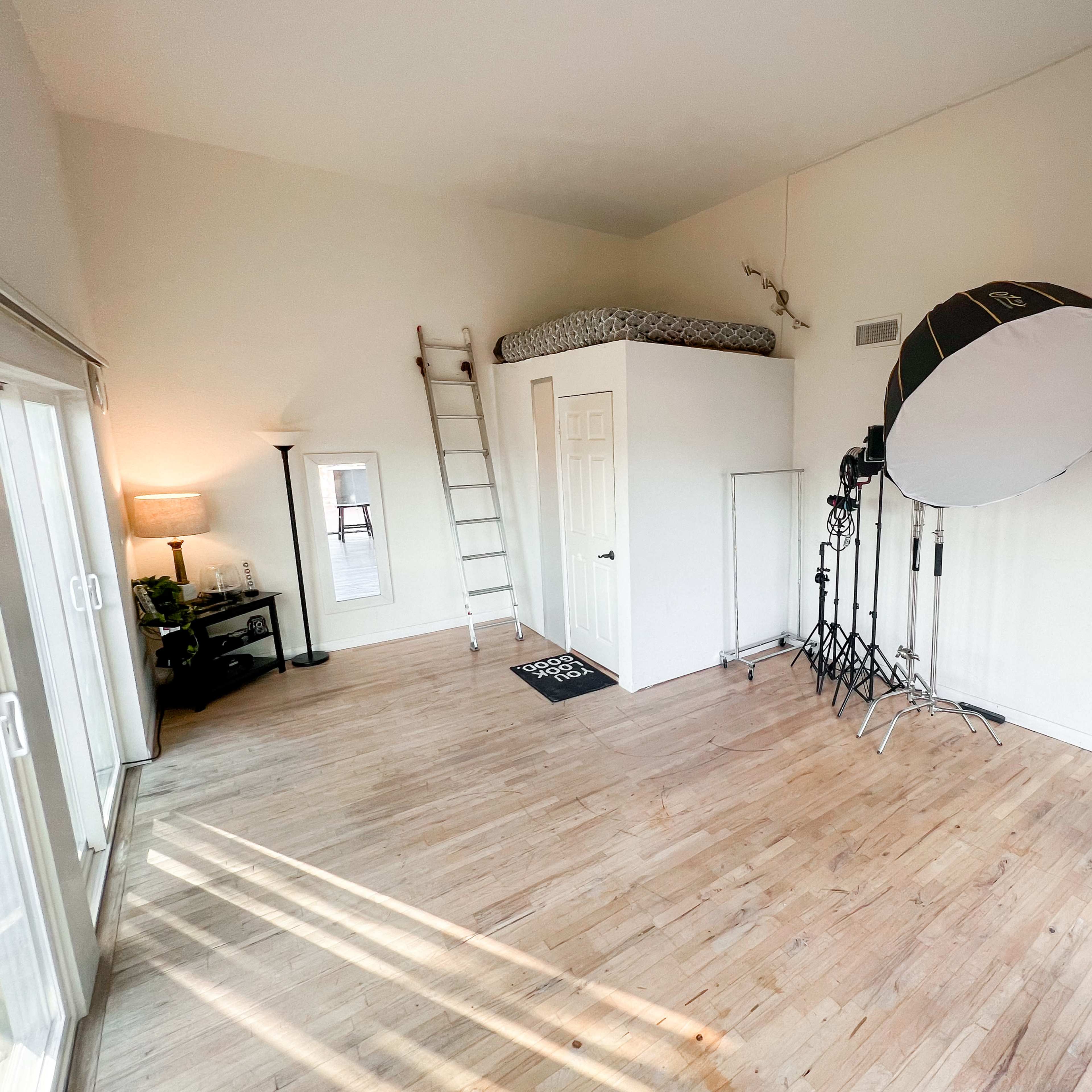 A spacious room with a bed on a lofted platform, a ladder, a large light stand with a softbox, and wooden floors.