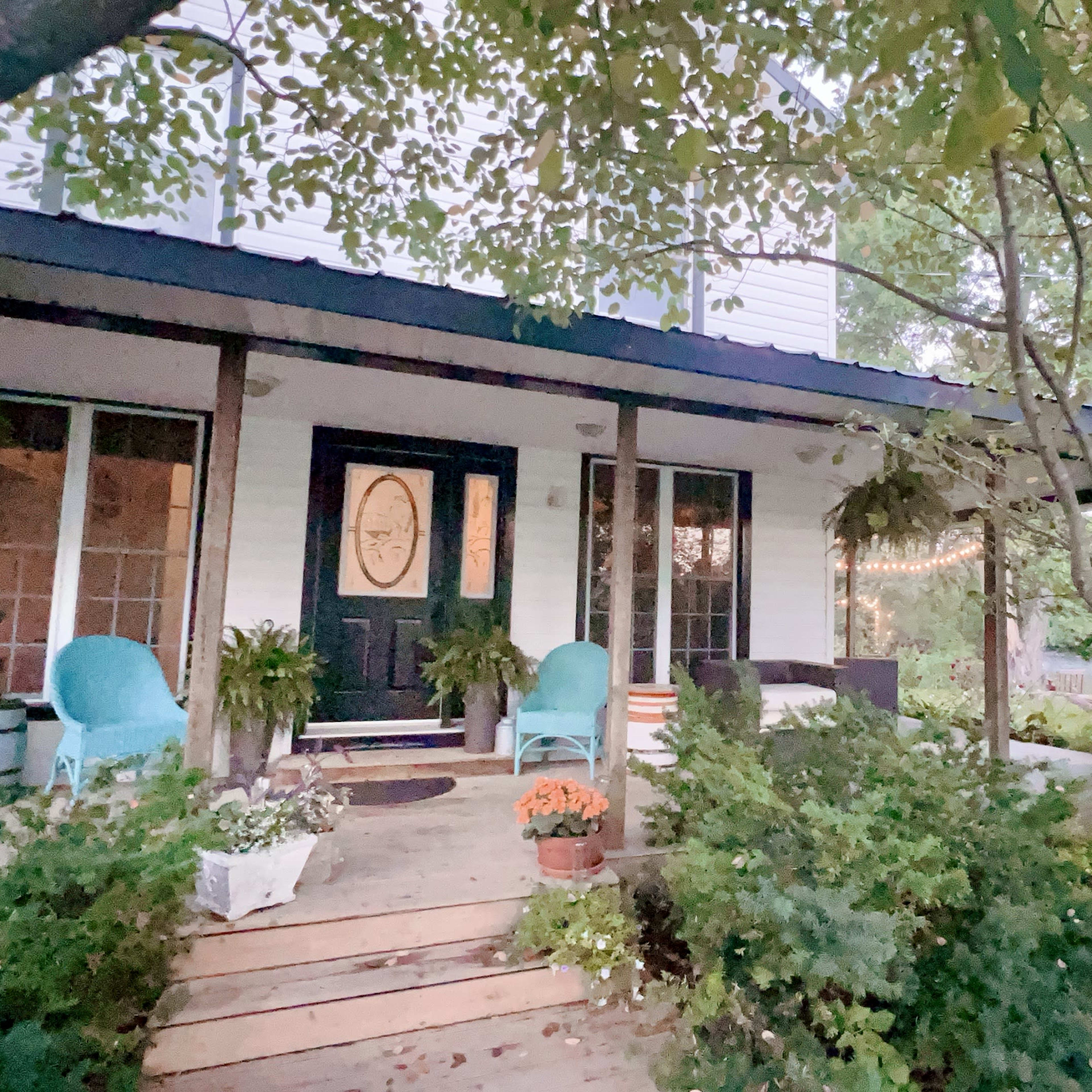 A two-story house features a front porch with two blue chairs, surrounded by greenery and potted plants.