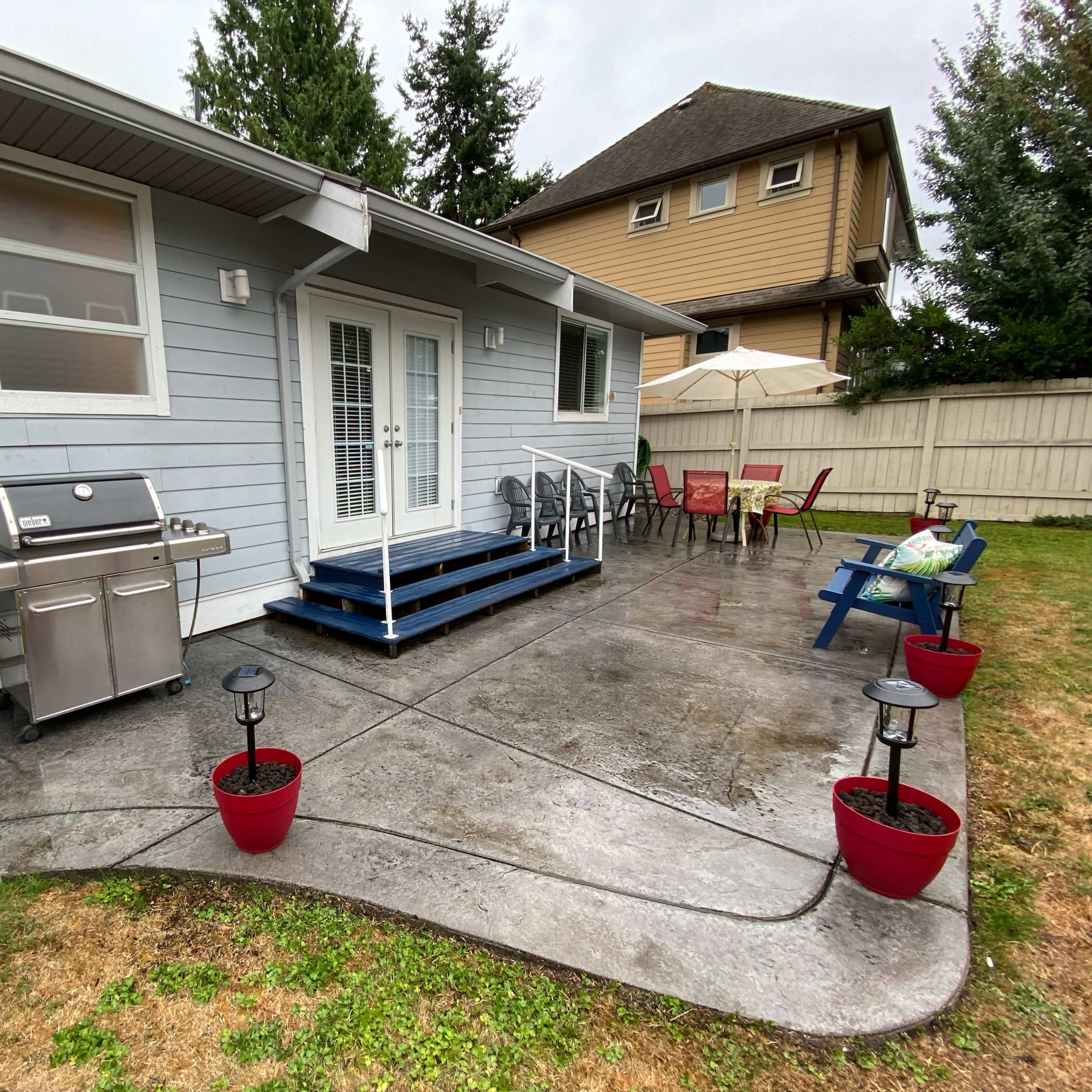 A gray house with a patio featuring a BBQ grill, outdoor seating, and potted plants on a rainy day.