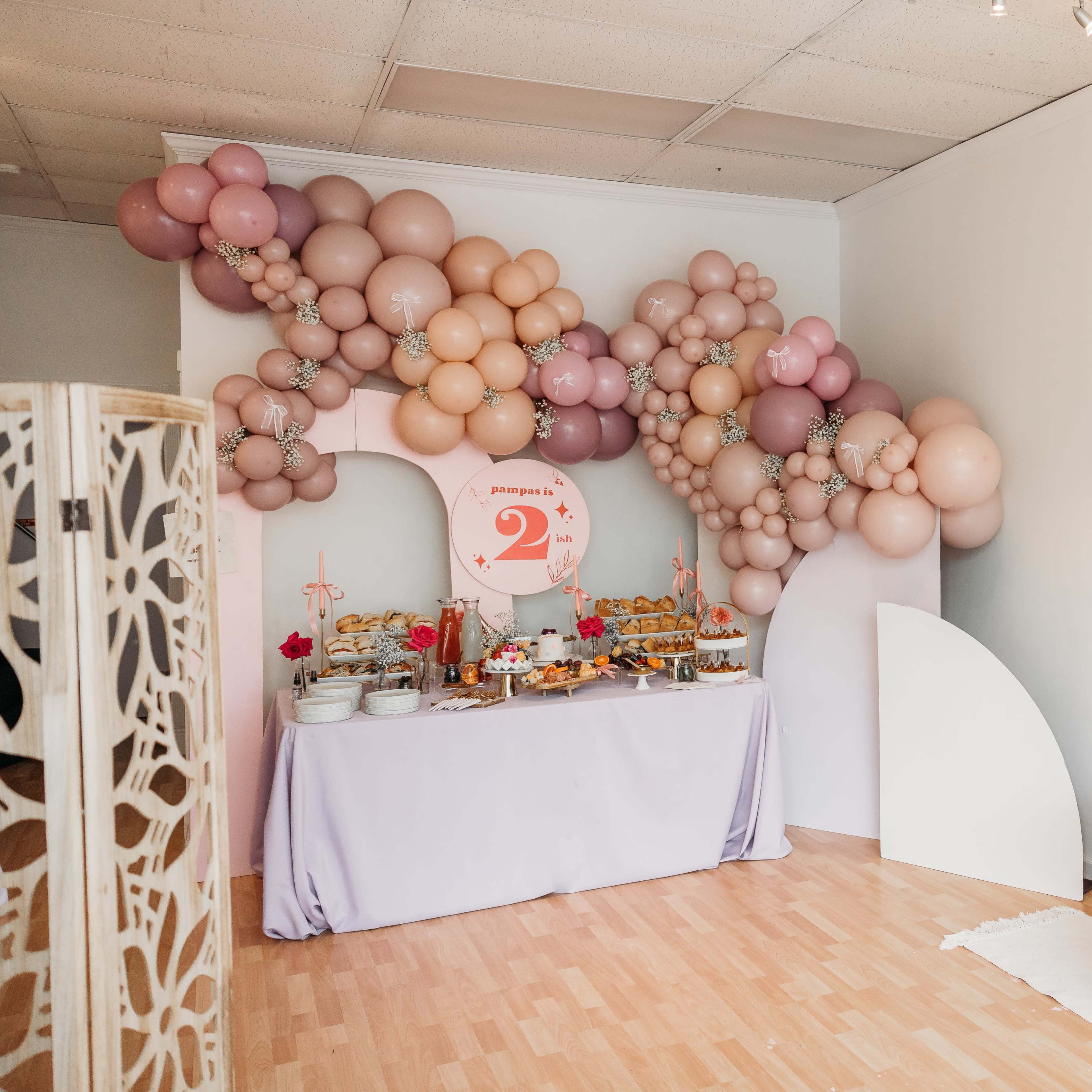 The scene features a decorated room with a table laden with food, a large balloon arch in shades of pink, and neatly displayed tote bags on the wall.