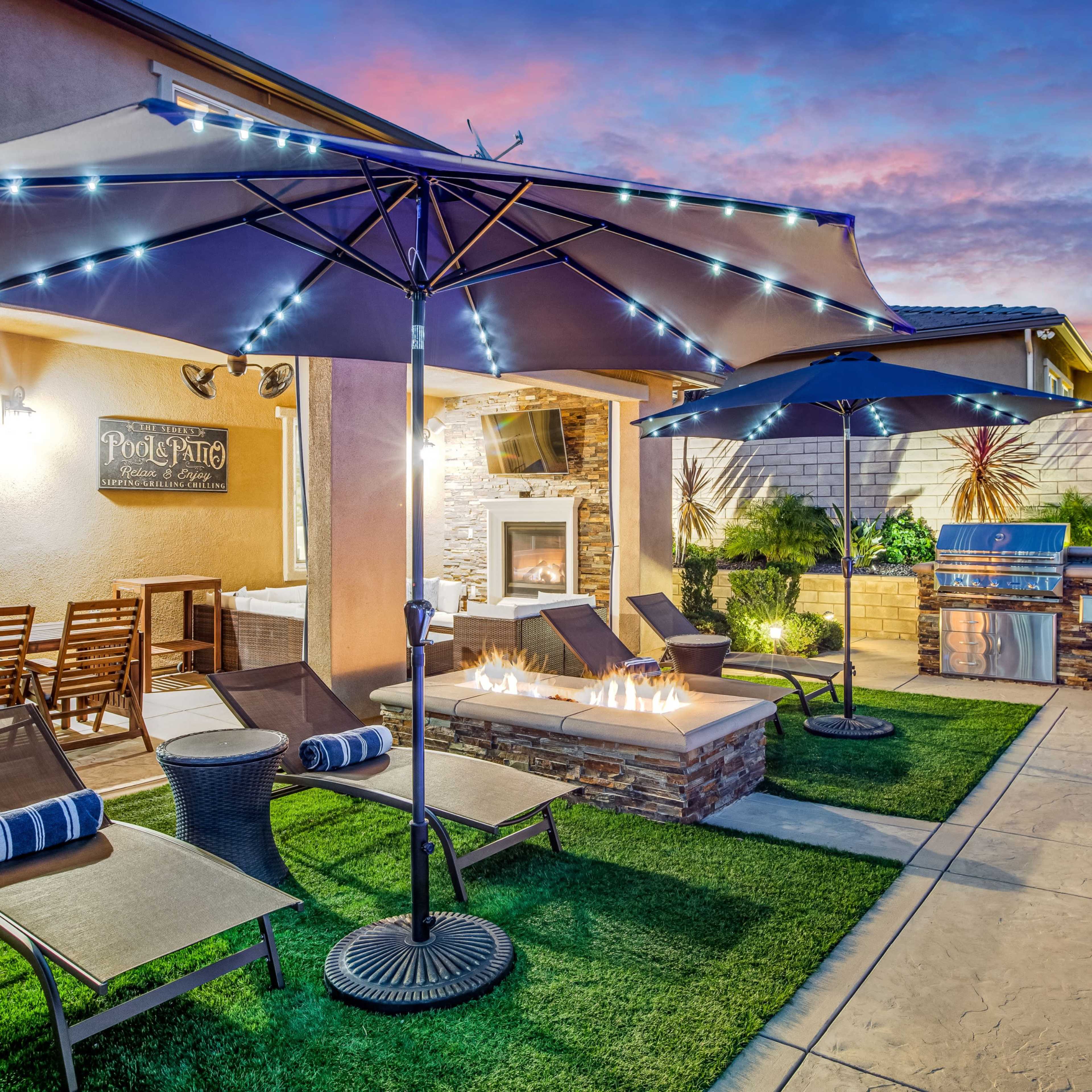 A backyard patio featuring two large umbrellas, lounge chairs, a fire pit, and an outdoor kitchen, set against a colorful sunset sky.