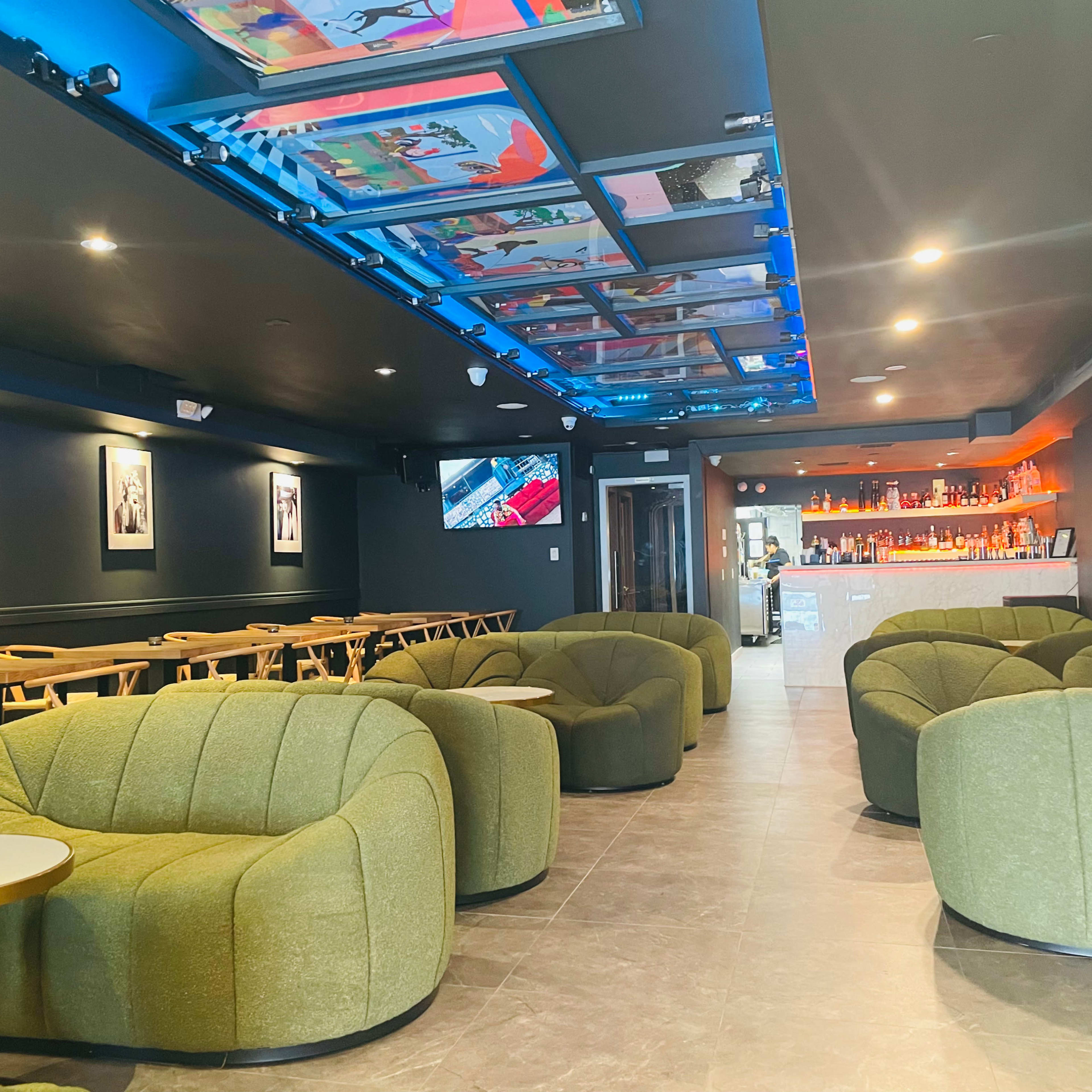 A modern lounge area with green upholstered chairs, a bar in the background, and colorful artwork displayed on the ceiling.