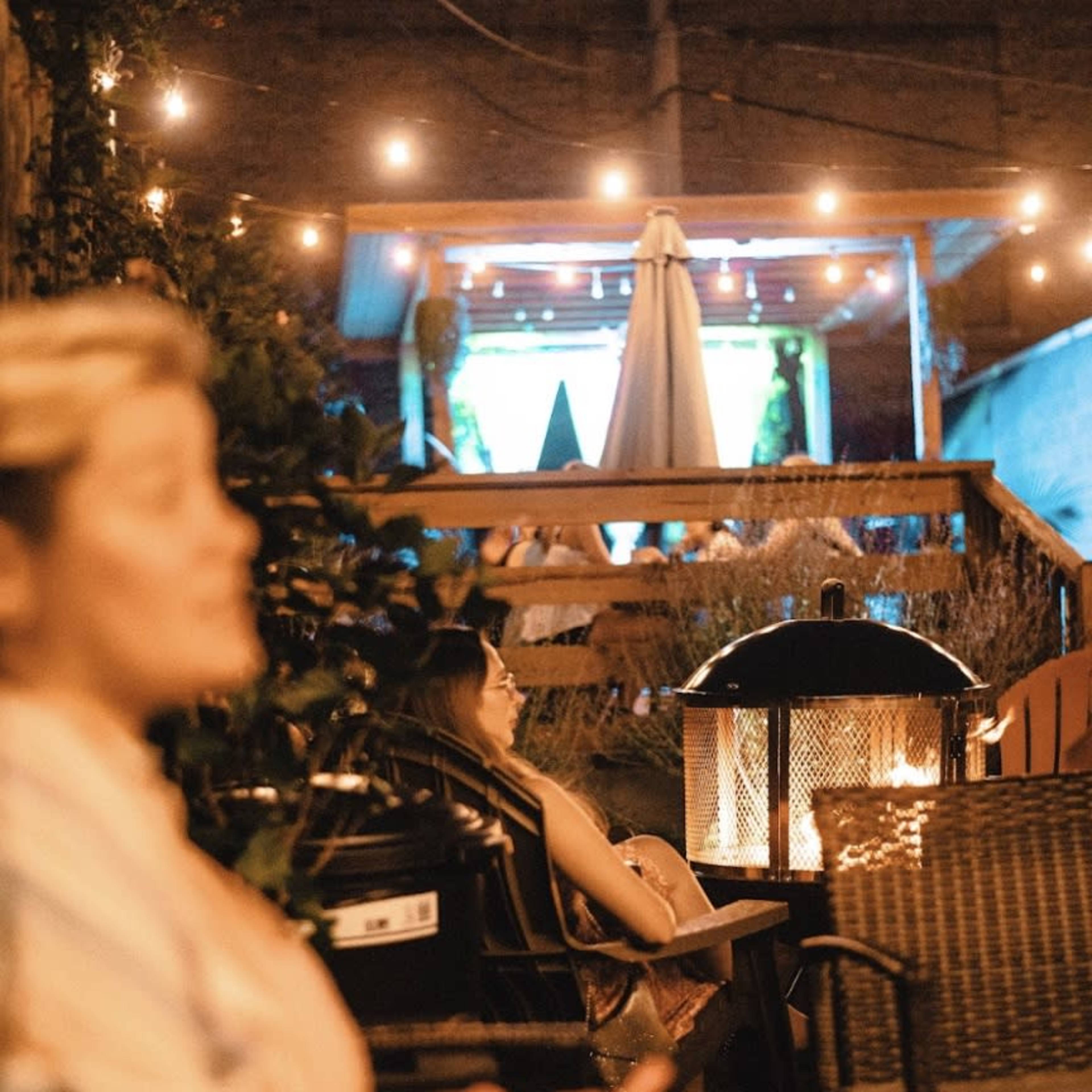 The scene depicts two people sitting in a cozy outdoor area with string lights illuminating the space and a fire pit in the foreground.