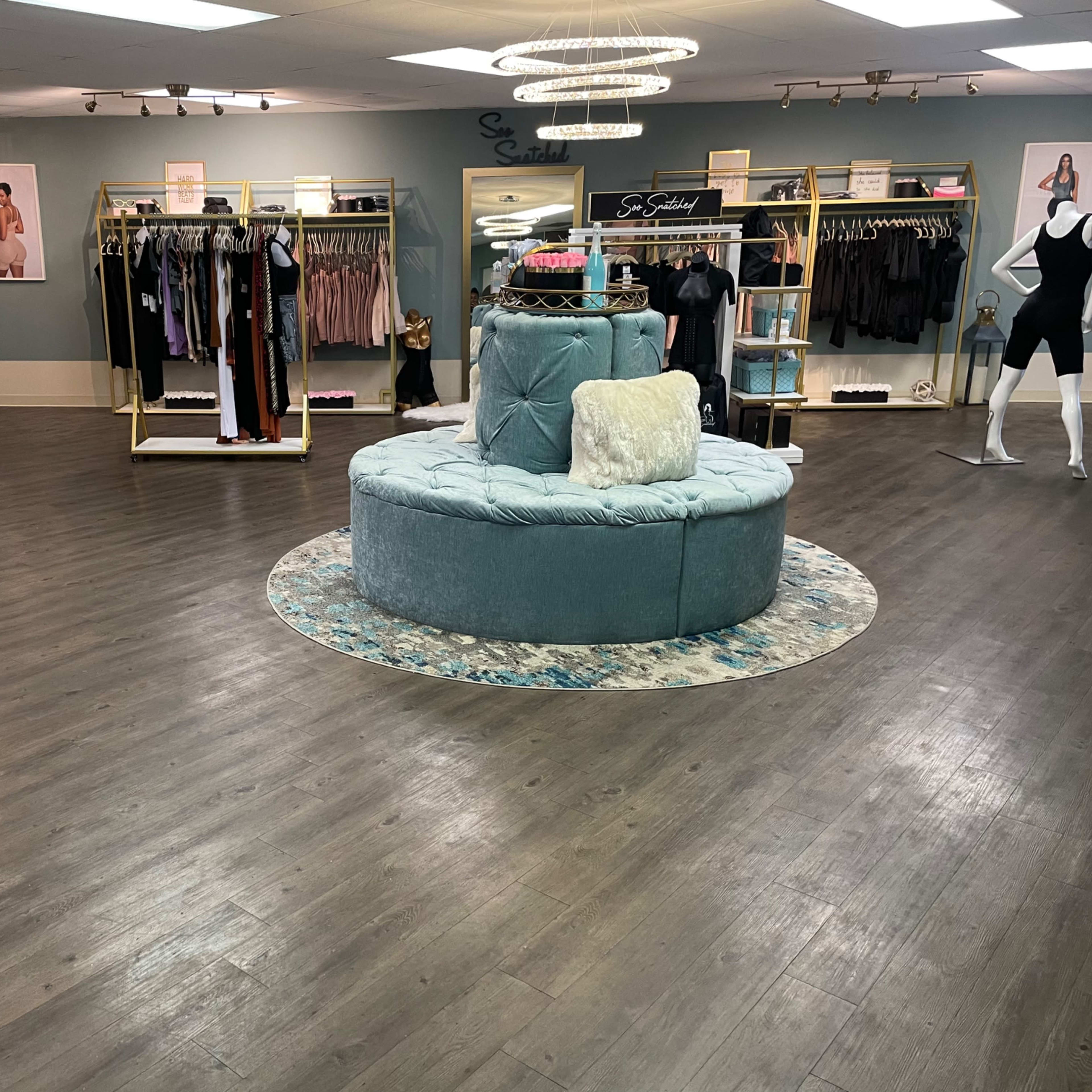 A spacious boutique with racks of clothing and a central circular seating area, featuring soft lighting and elegant decor.