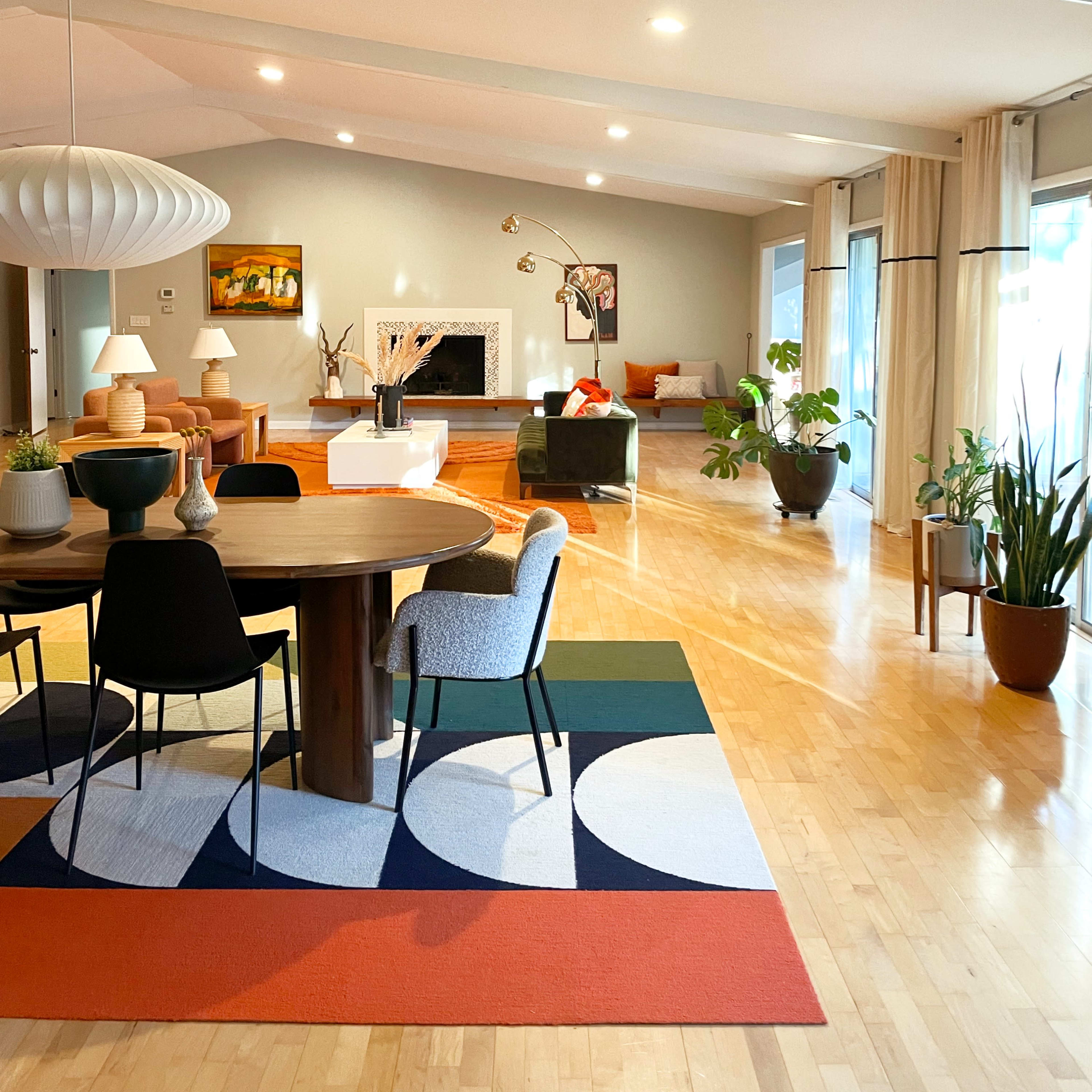 A spacious living area with a round dining table at the center, surrounded by modern chairs and a large area rug, featuring large windows and various indoor plants.