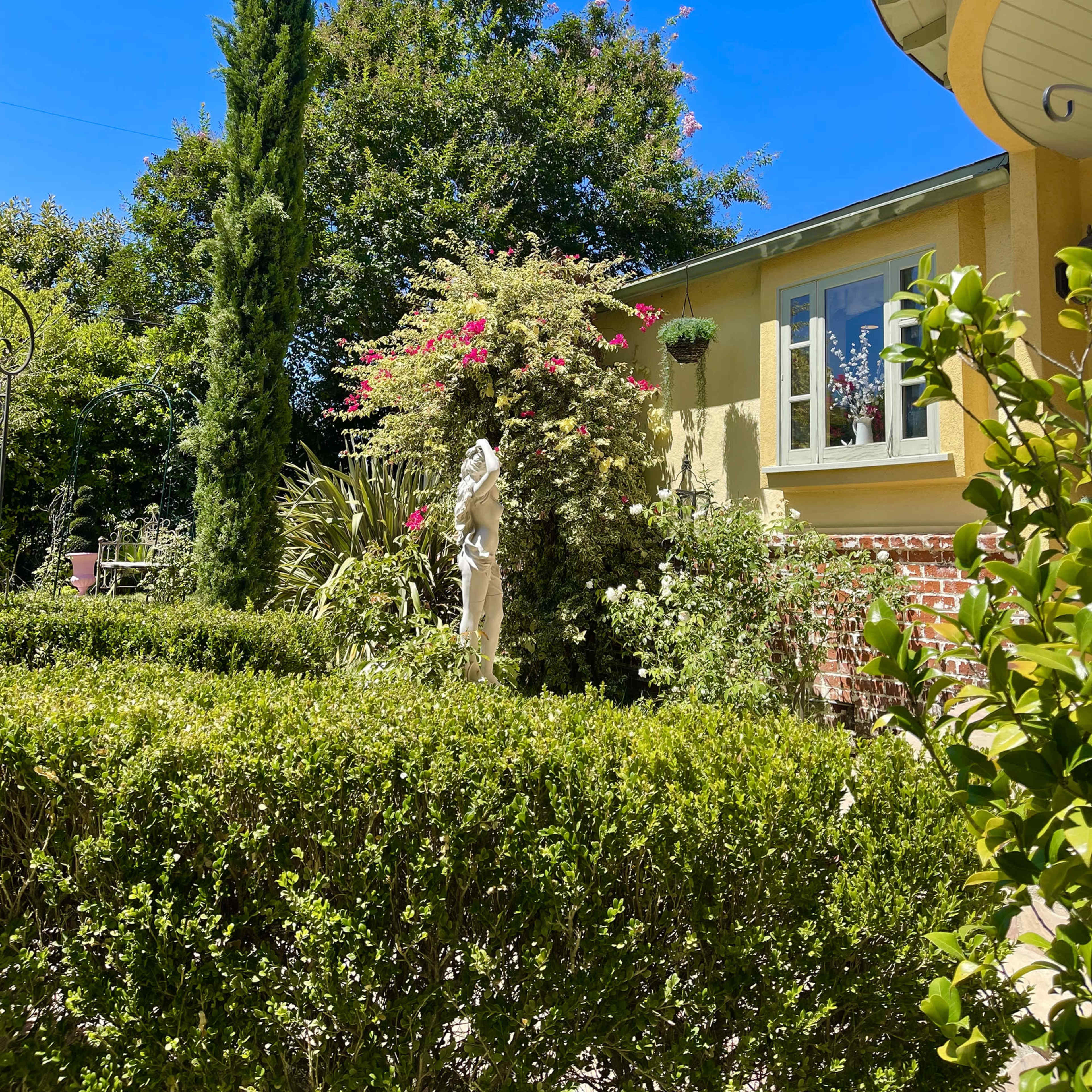 A well-maintained garden features a tall cypress tree, vibrant flowering plants, and a statue beside a house with a yellow exterior.