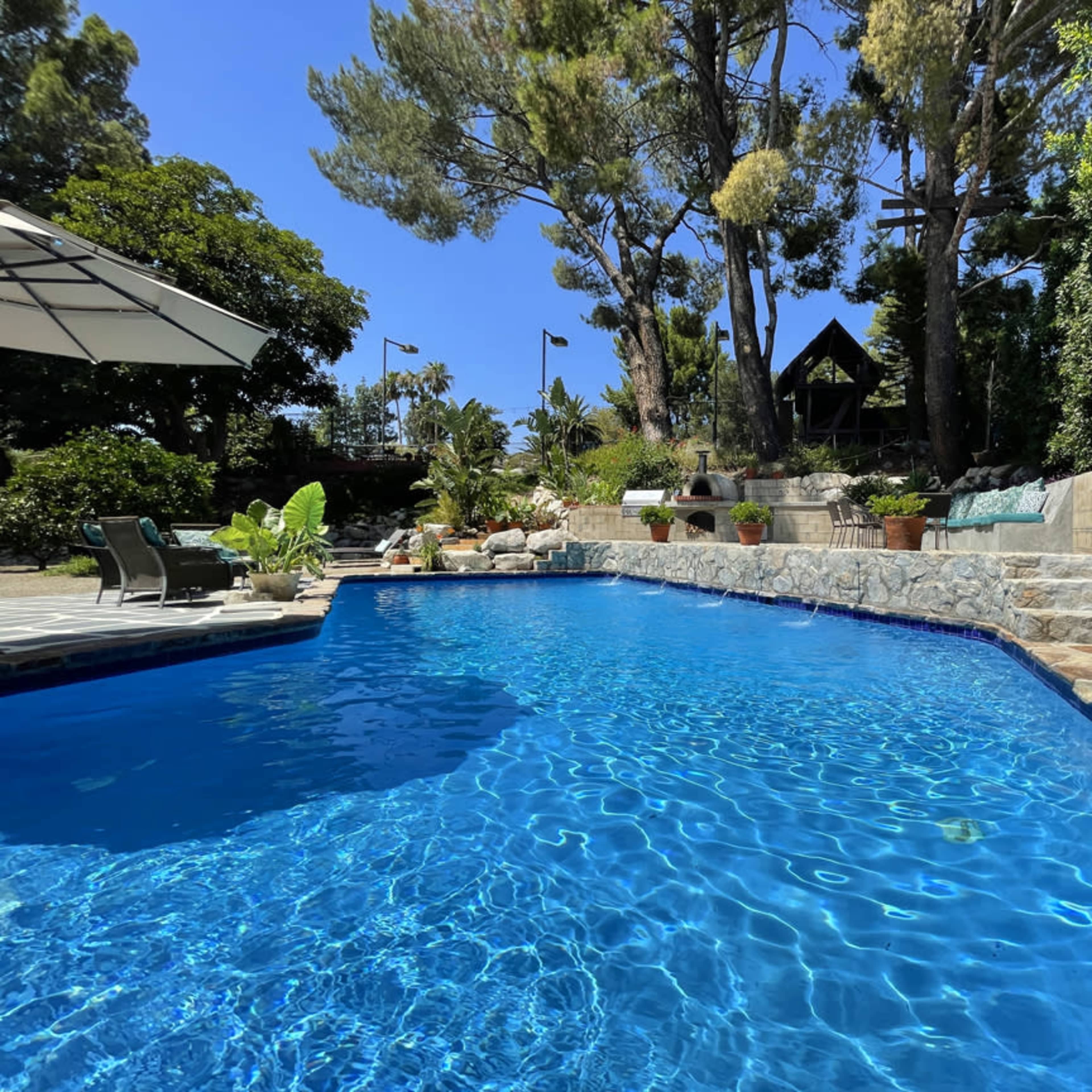 A clear blue swimming pool surrounded by stone steps, greenery, and tall trees under a bright sky.