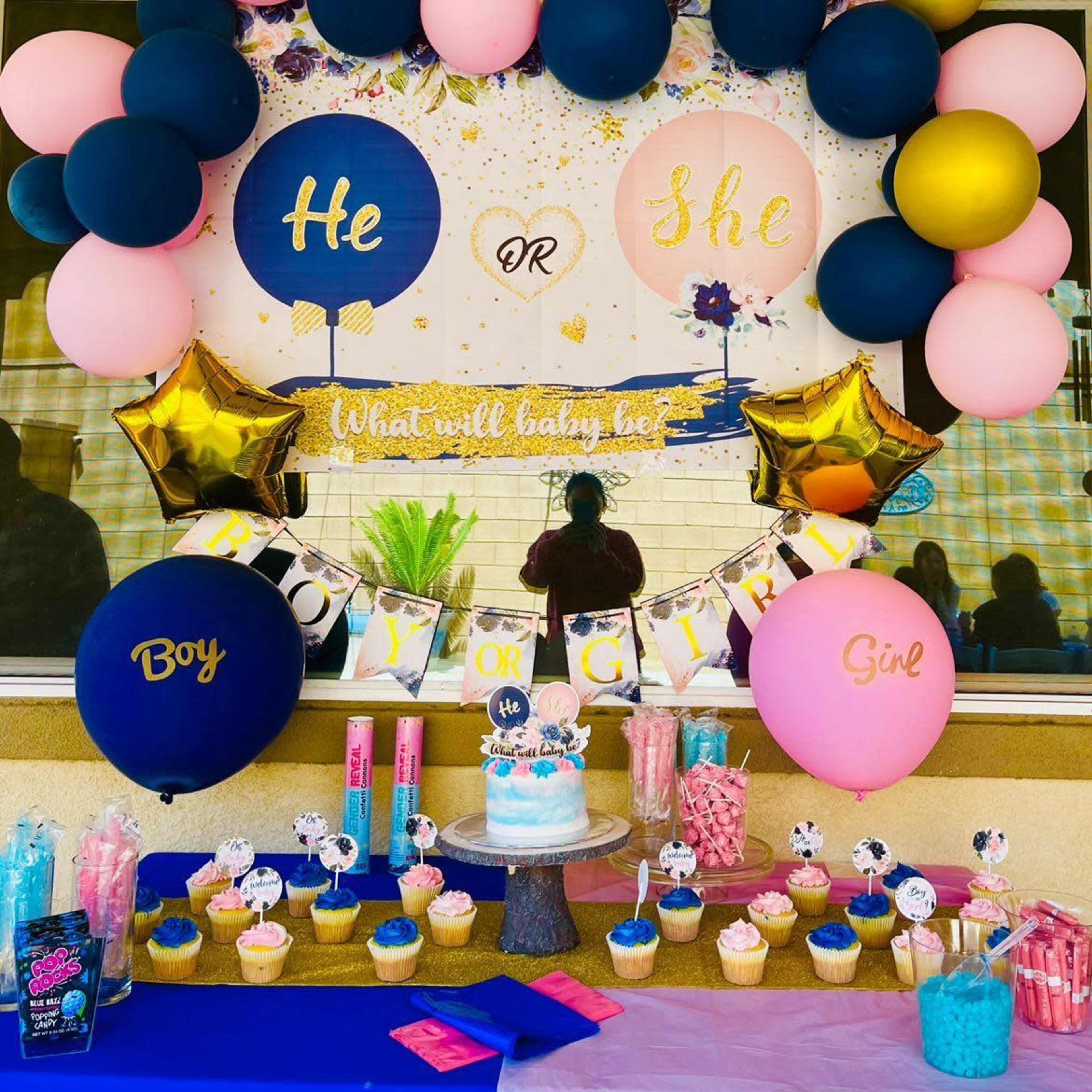 A decorated table displays a cake, cupcakes, candy, and balloons for a gender reveal party.