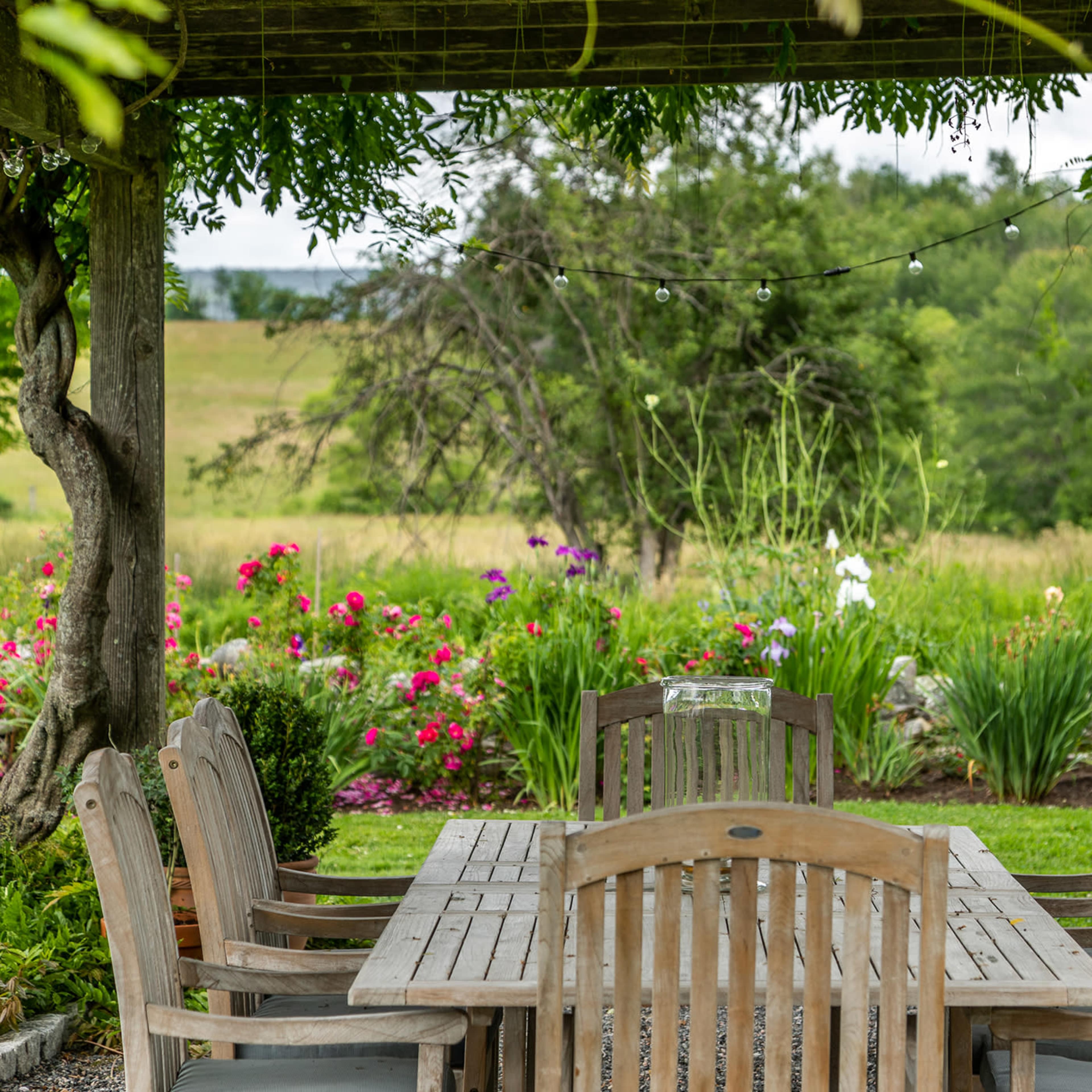 A wooden dining table surrounded by chairs is set under a pergola with string lights, overlooking a vibrant garden and open fields.