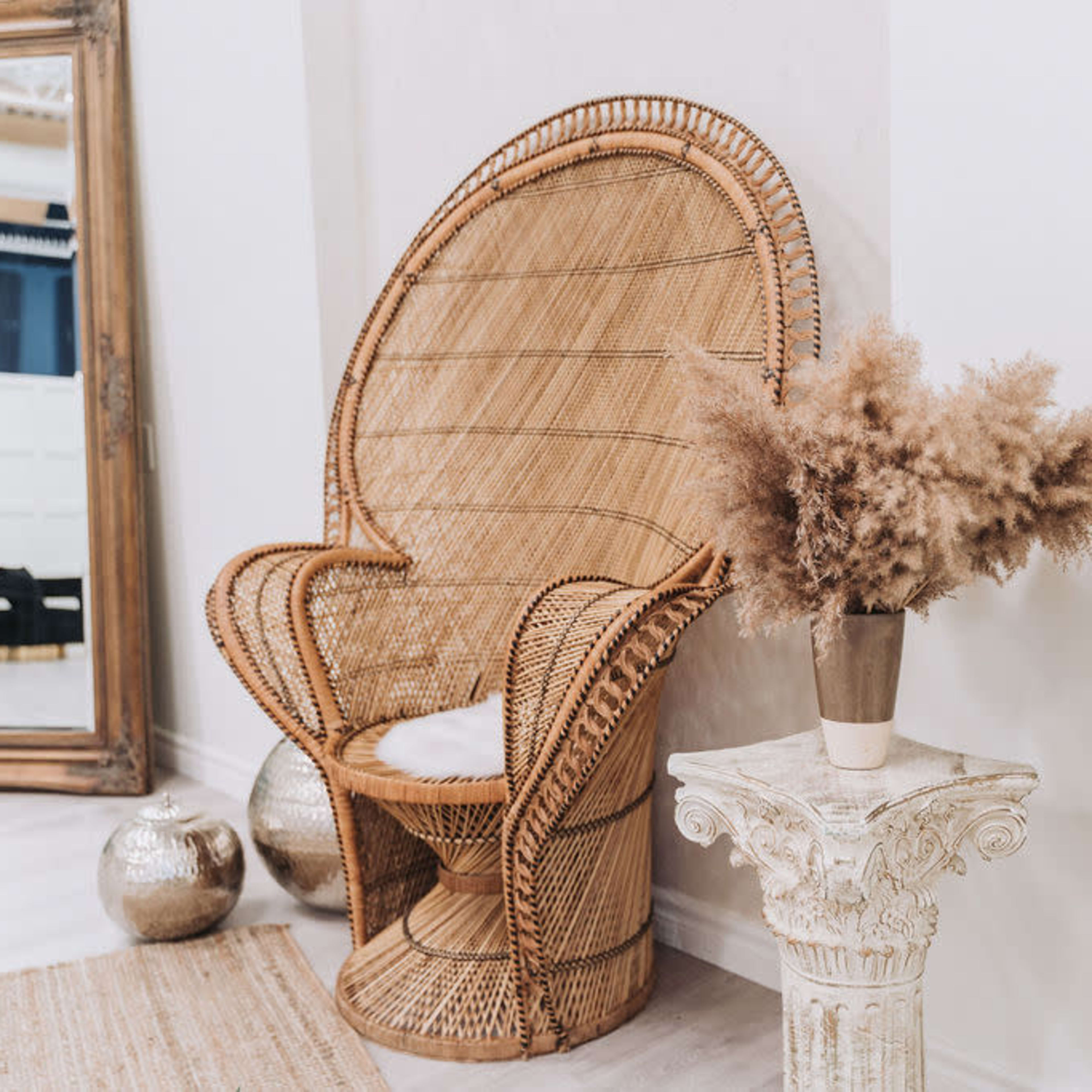 A large, woven rattan chair is positioned next to a decorative column with a vase of dried flowers, against a light-colored wall and a large mirror in the background.
