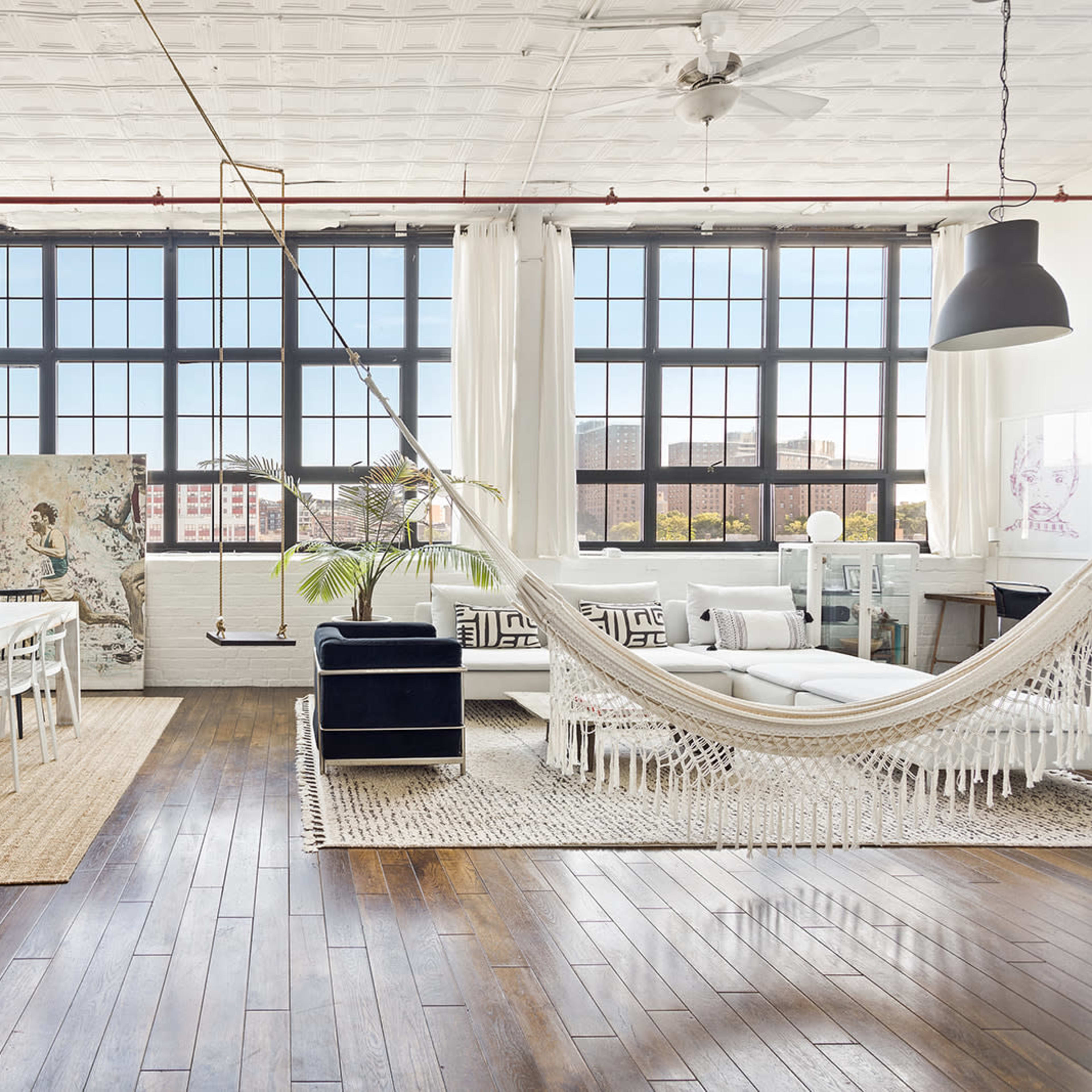 A spacious, well-lit room features a dining area with a table and chairs, a hammock, and a seating area with a sofa, large windows, and hardwood flooring.