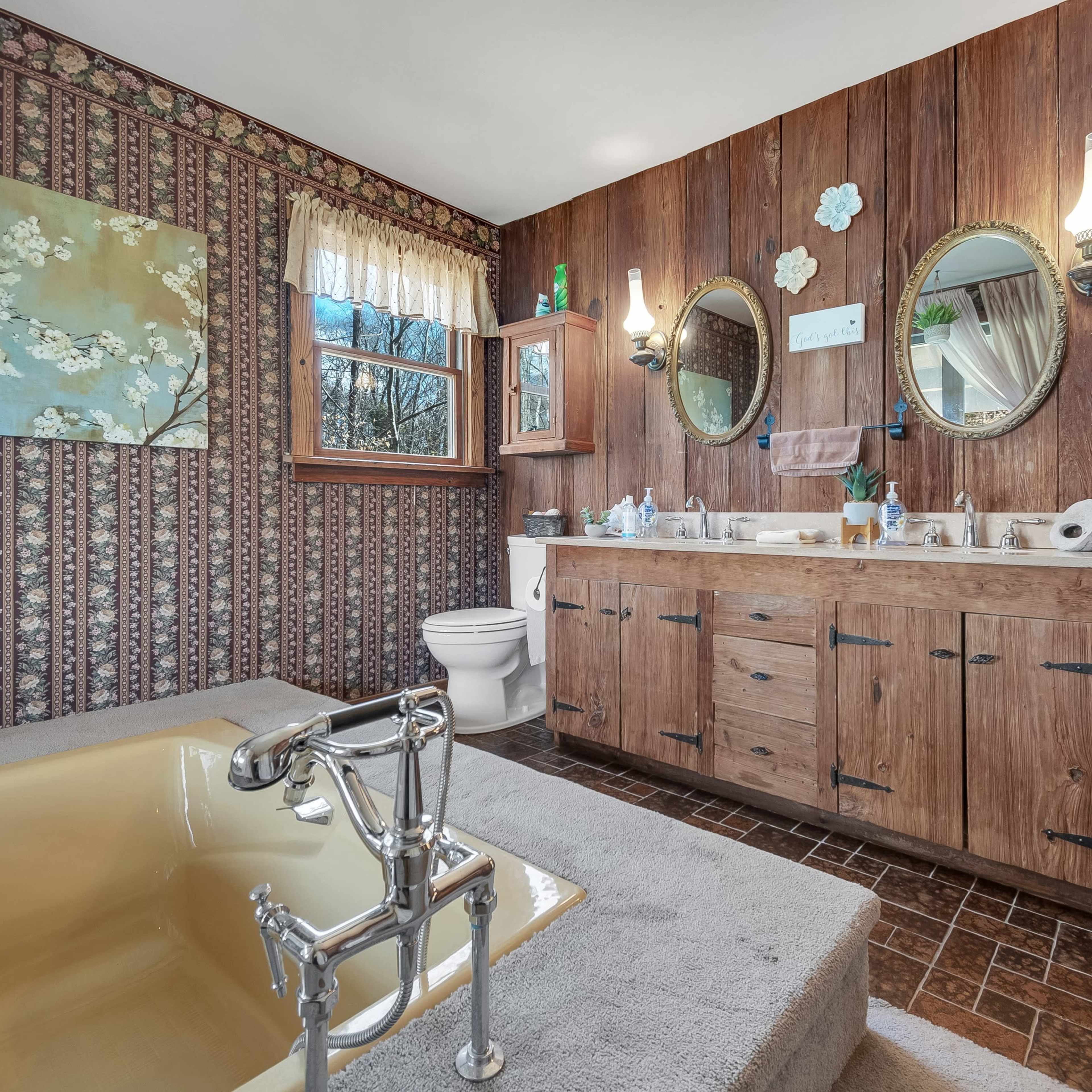 A bathroom with wood-paneled walls, a freestanding yellow bathtub, a double vanity with mirrors, and floral wallpaper.