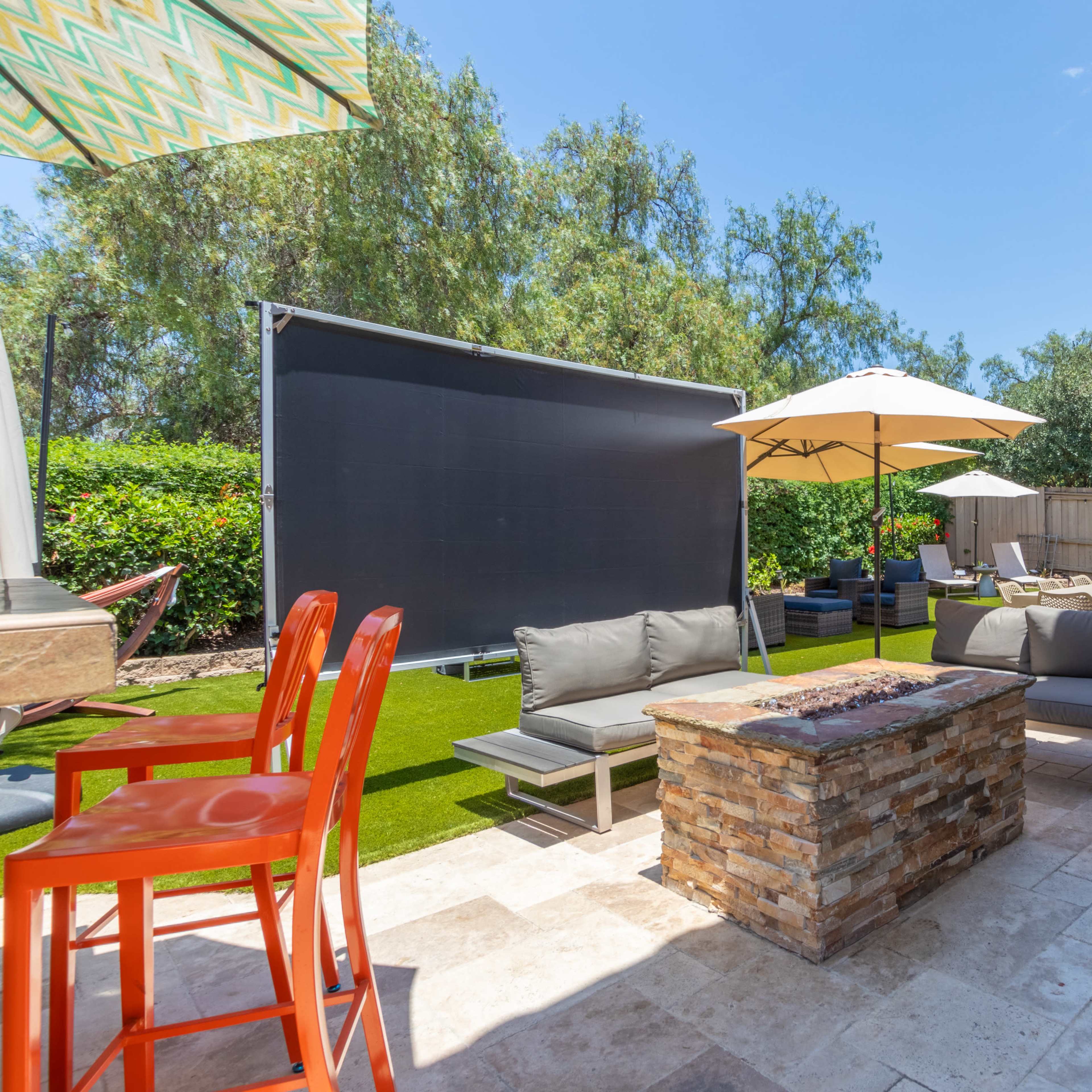 The outdoor area features a stone fire pit surrounded by modern seating, with umbrellas providing shade and a large screen set up in the background.